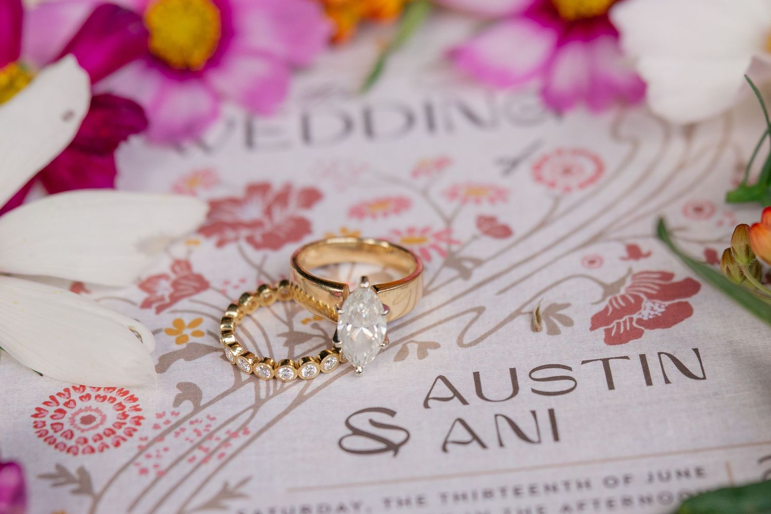 Gold wedding rings rest on a floral wedding invitation for Austin and Ani, surrounded by colorful blooms.