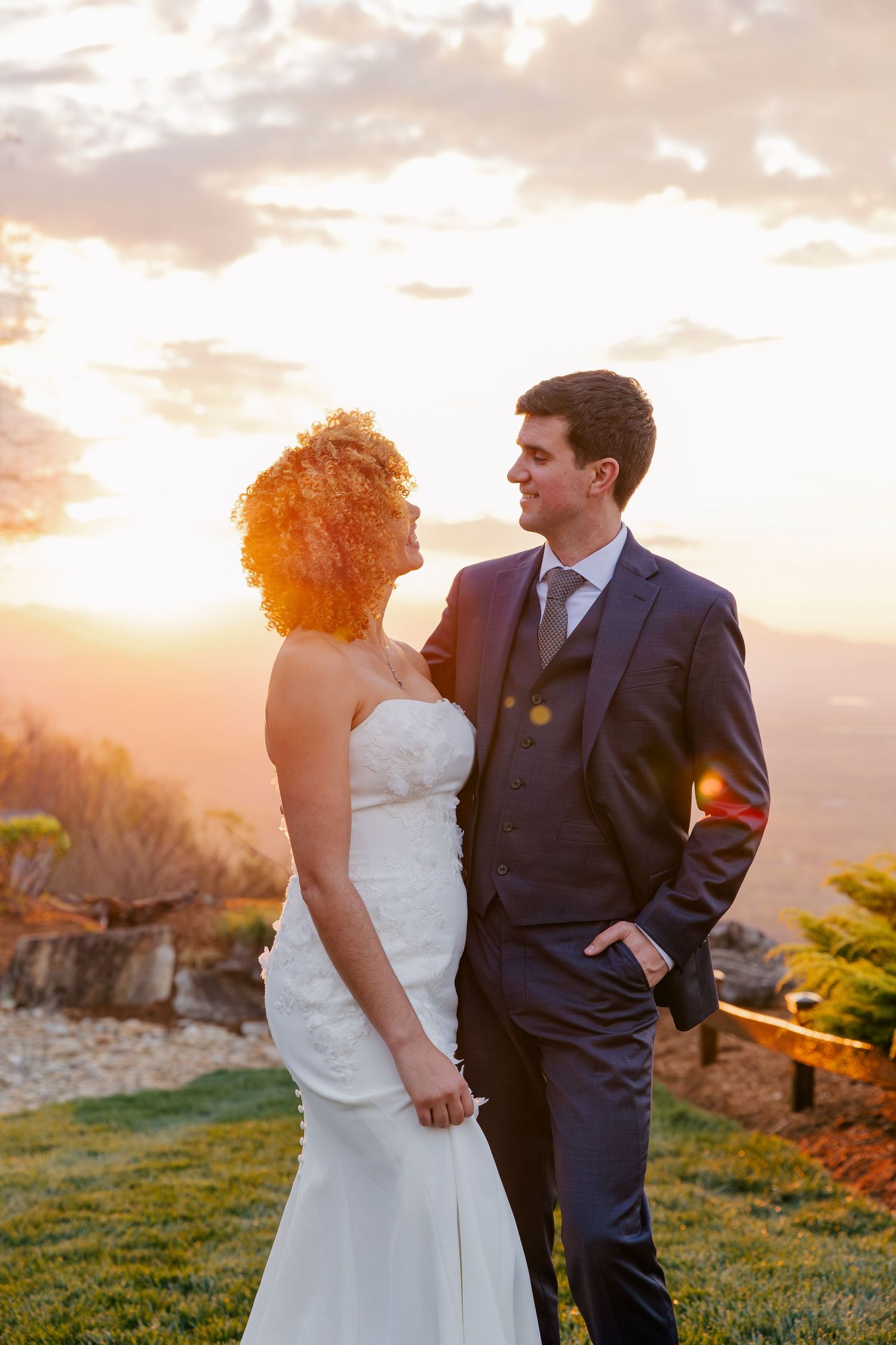 Newlyweds pose romantically at golden hour sunset, bride in strapless white gown and groom in navy three-piece suit.
