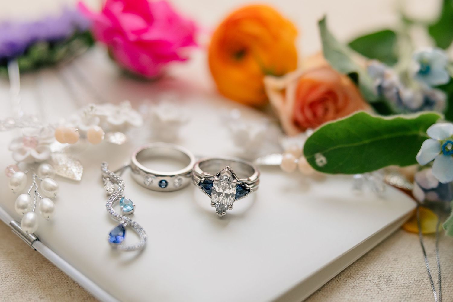 Elegant bridal jewelry flat lay featuring diamond engagement ring, sapphire wedding band, and colorful blooms.