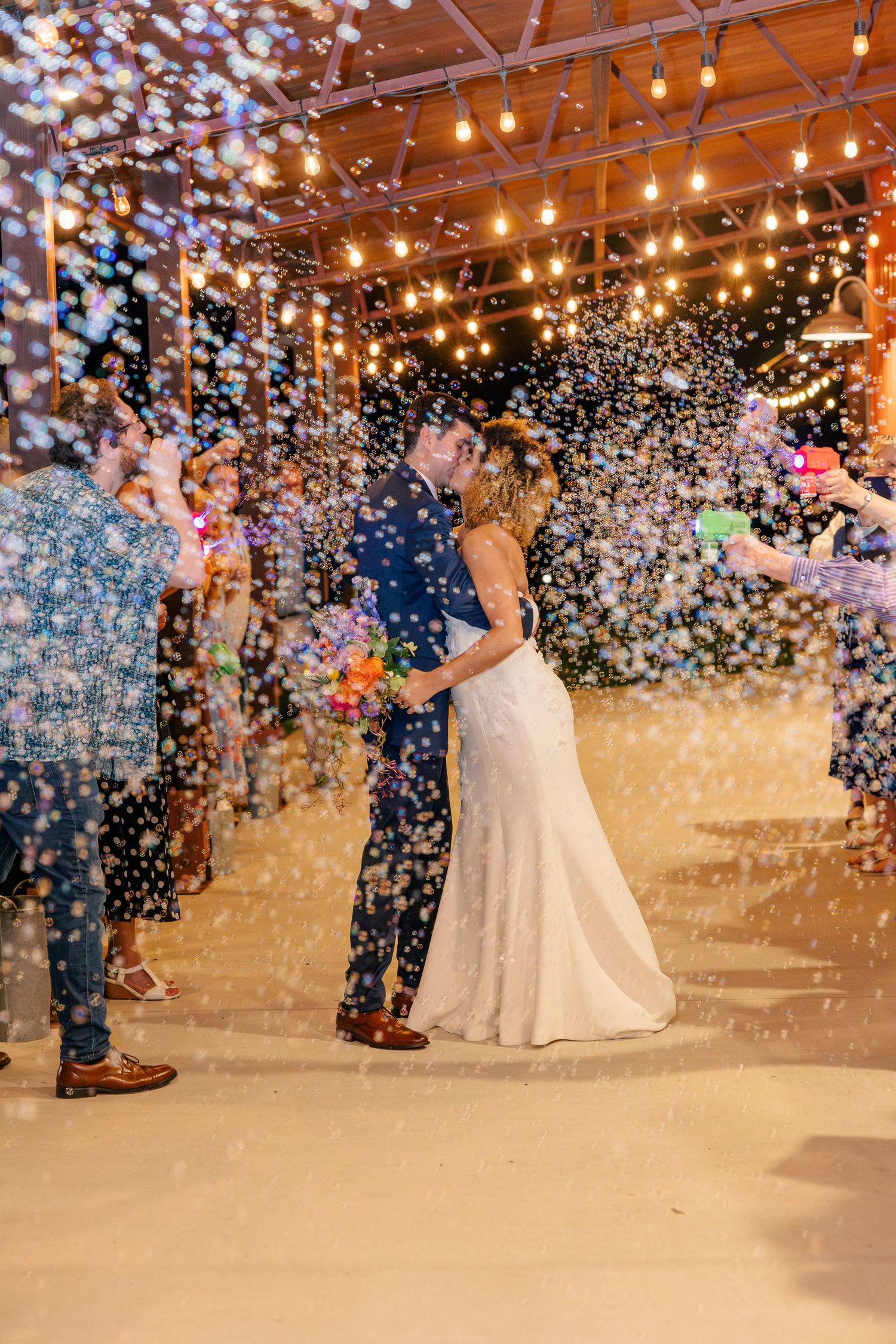 Newlyweds sharing first dance surrounded by bubbles under warm string lights at wedding reception.