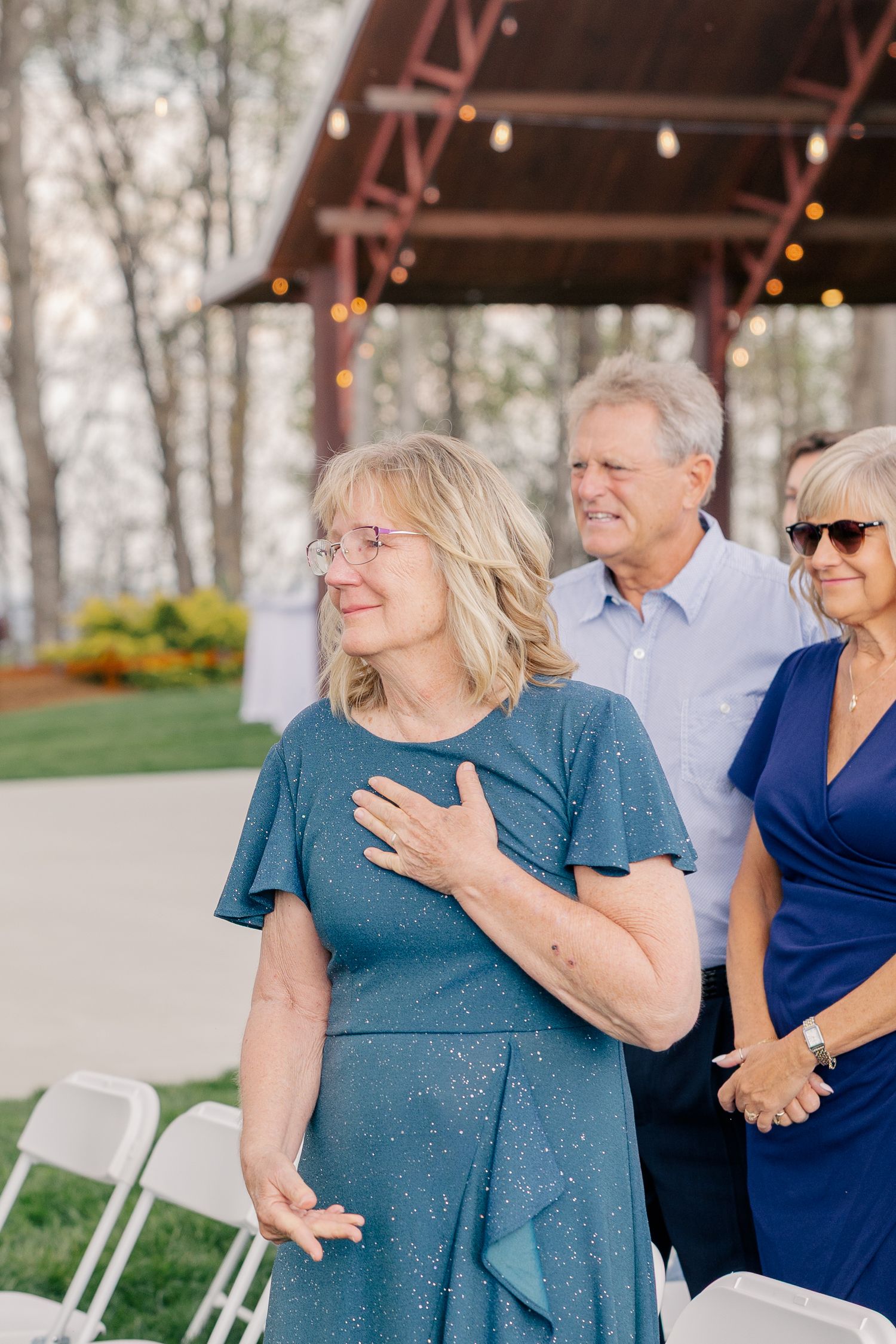 Emotional woman in teal dress places hand on chest during an outdoor wedding ceremony with string lights.
