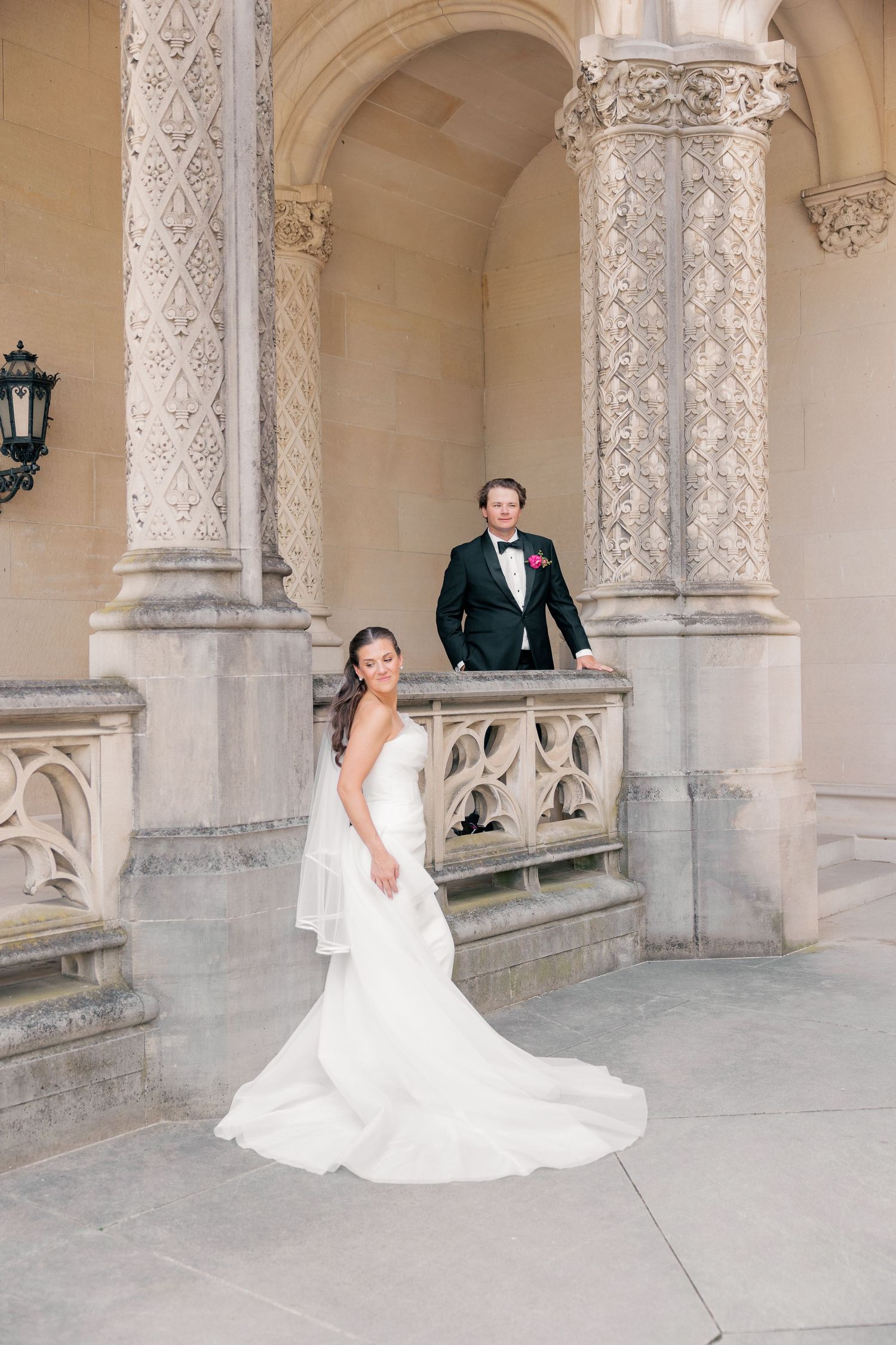 Bride in white gown and groom in black tuxedo pose at ornate stone archway columns for elegant wedding portrait.