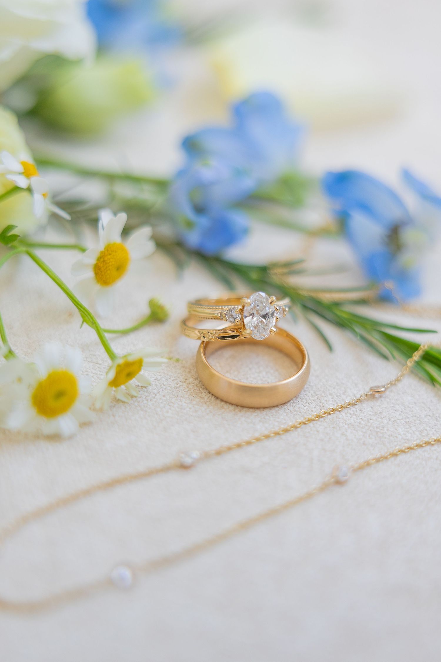 Gold wedding rings and engagement ring with oval diamond beside daisies and blue flowers on white surface.
