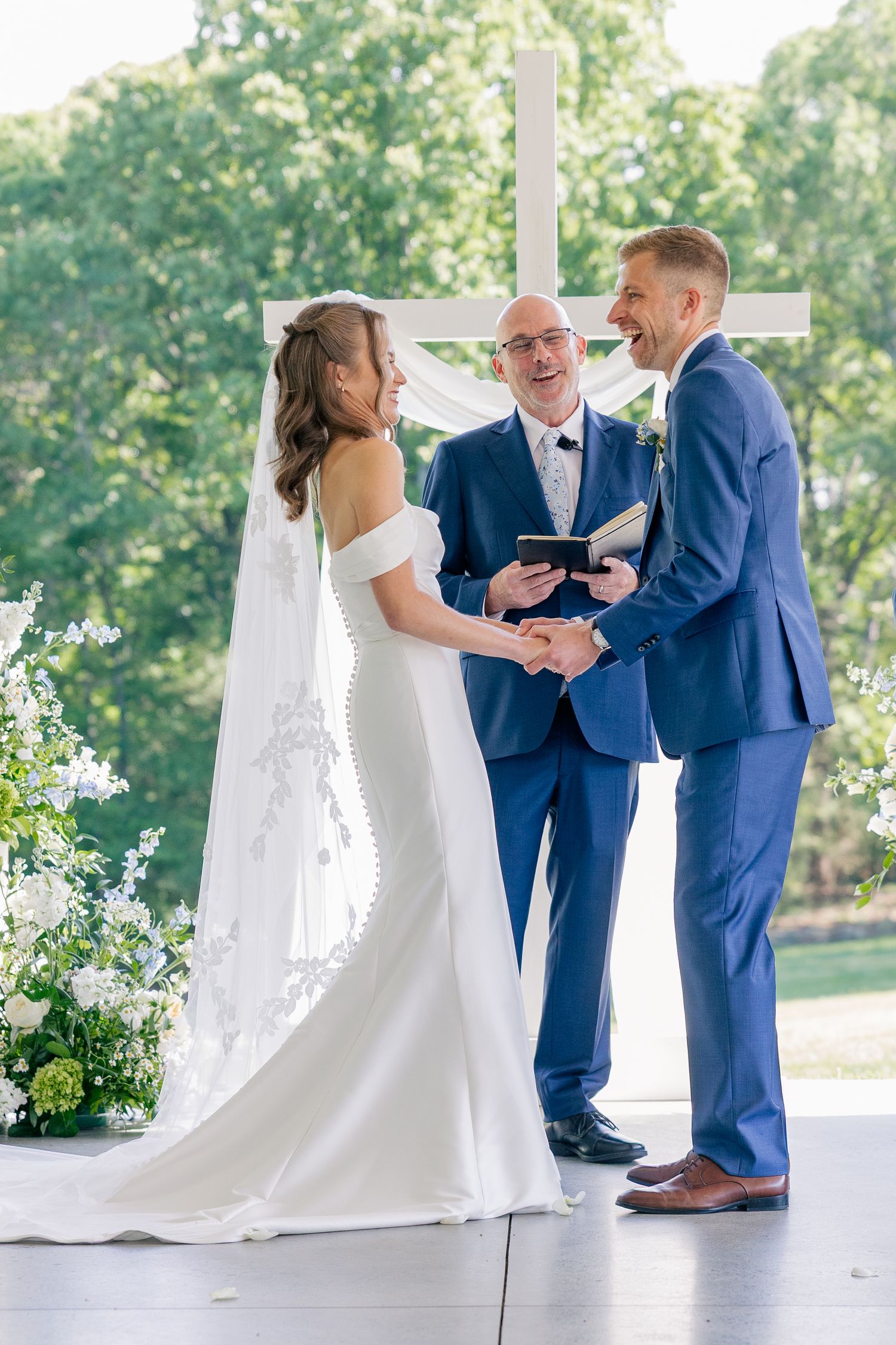 Bride and groom hold hands laughing at outdoor wedding ceremony with officiant before white cross.