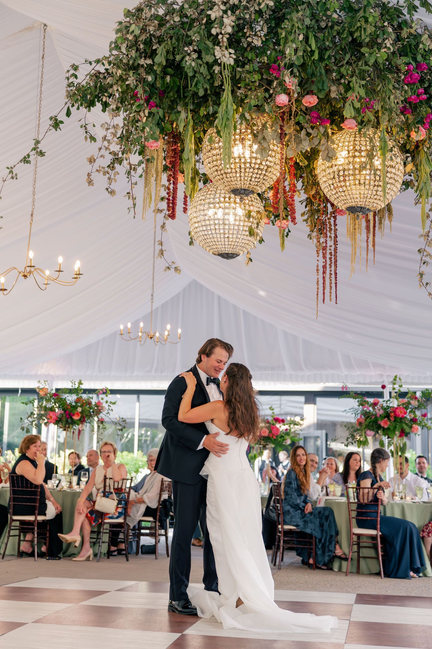 Bride and groom share first dance under lush floral chandelier installation at elegant tented wedding reception.