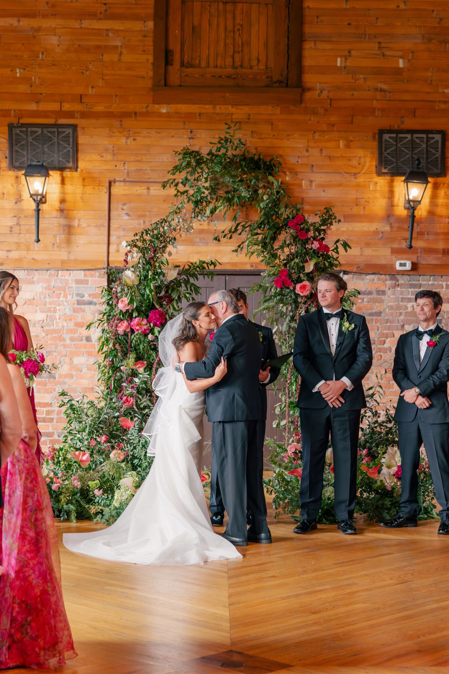Bride and groom share first kiss at floral arch altar in rustic barn wedding ceremony.