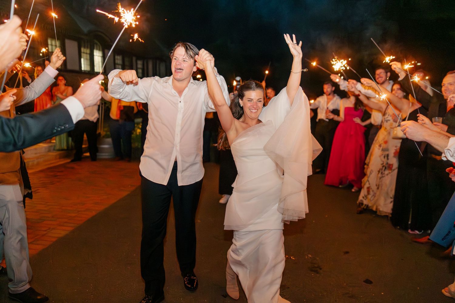 Bride and groom celebrate with sparkler exit surrounded by cheering wedding guests at night.