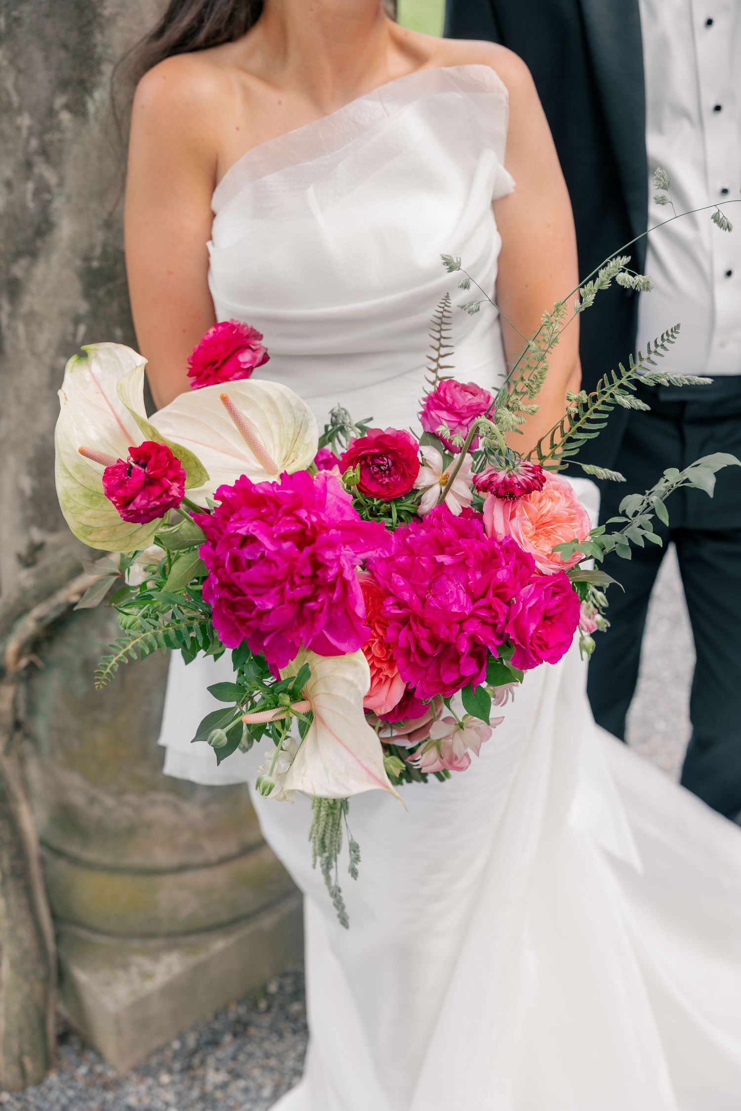 Bride in white one-shoulder gown holding vibrant fuchsia peony and anthurium wedding bouquet with groom.