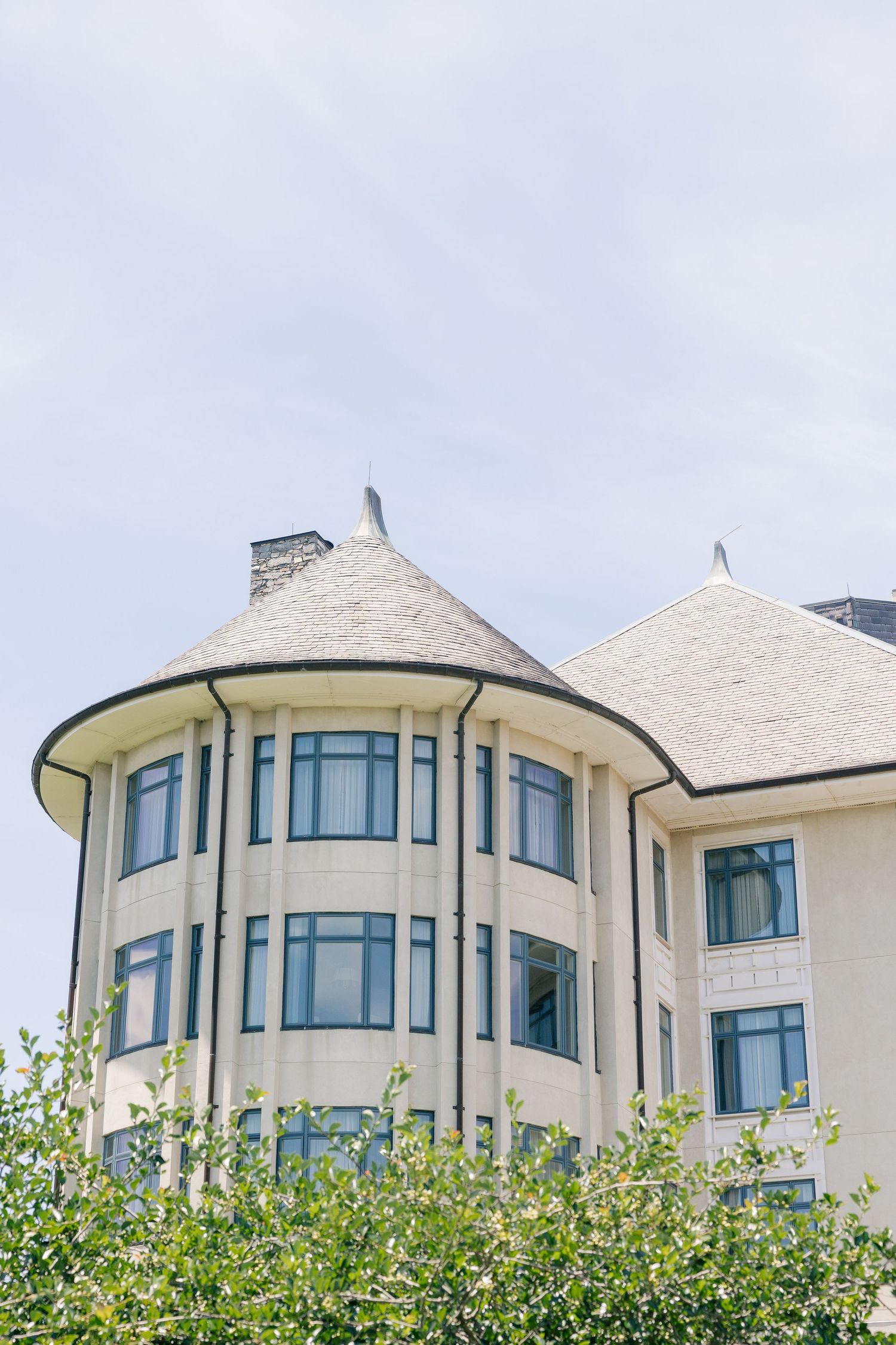 Elegant building with conical turret roofs and large windows surrounded by green trees.