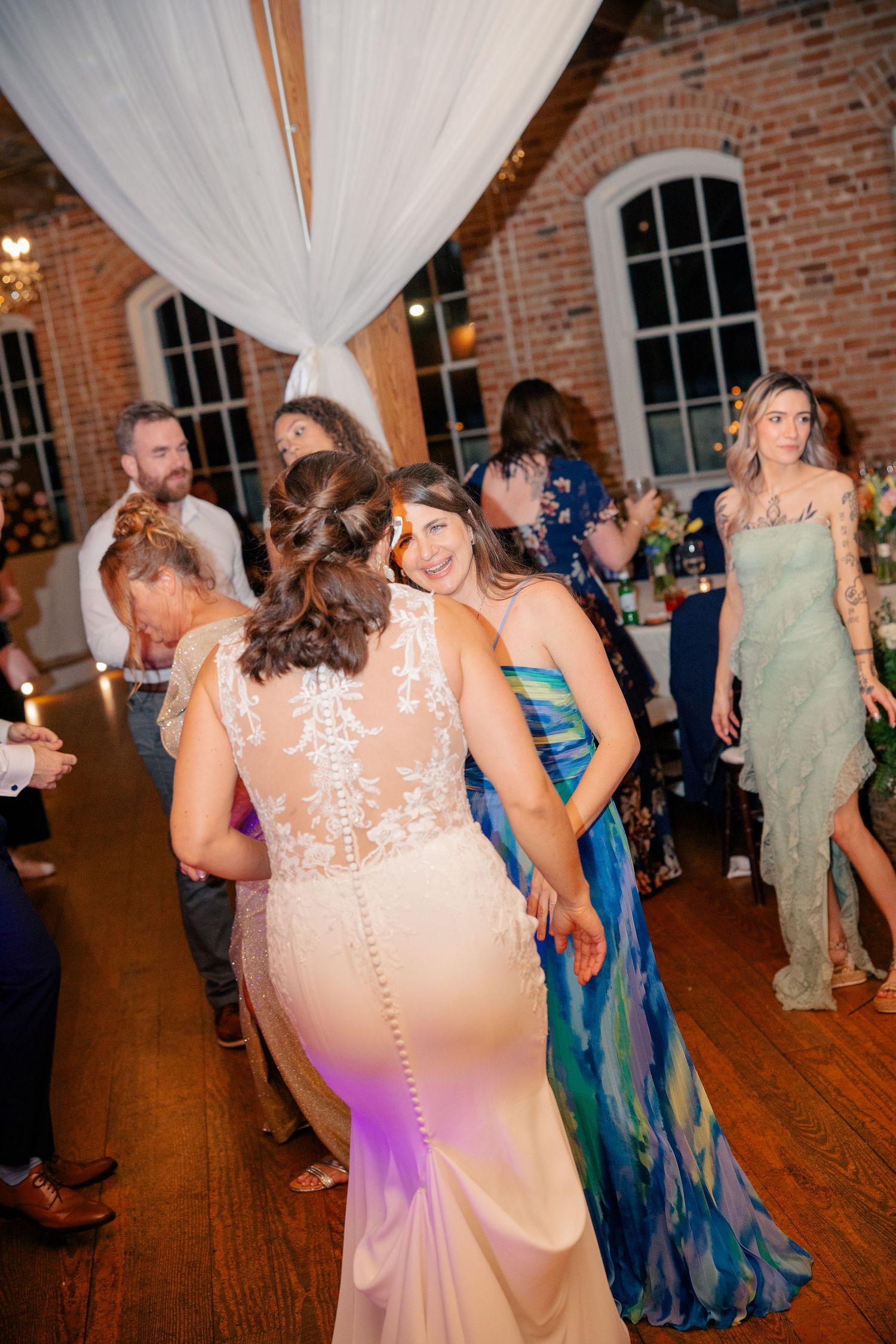 Bride in lace back gown dances joyfully with guests on decorated wedding reception dance floor.
