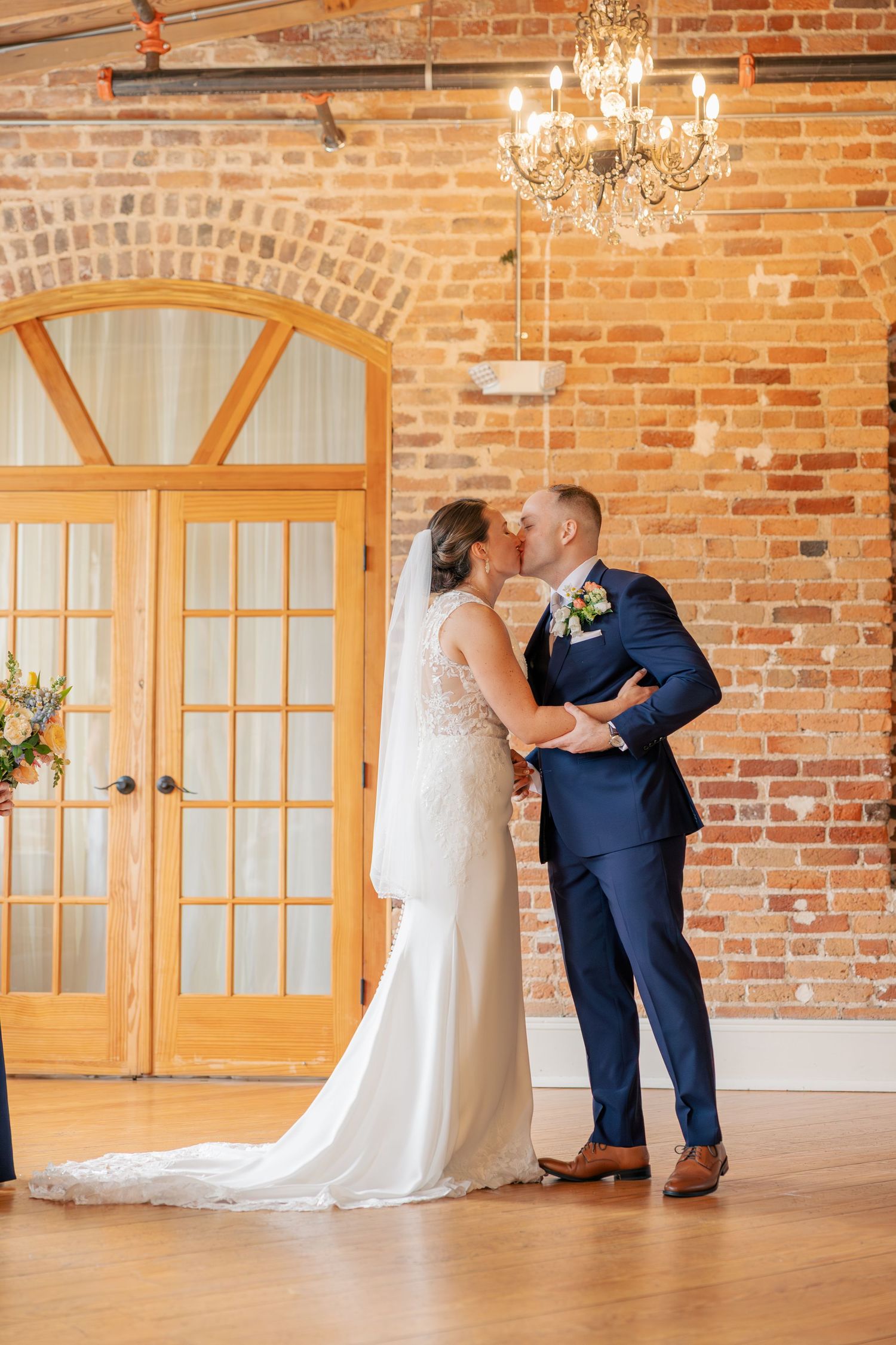 Bride and groom share first kiss at altar inside a rustic brick wall venue with chandelier and French doors.
