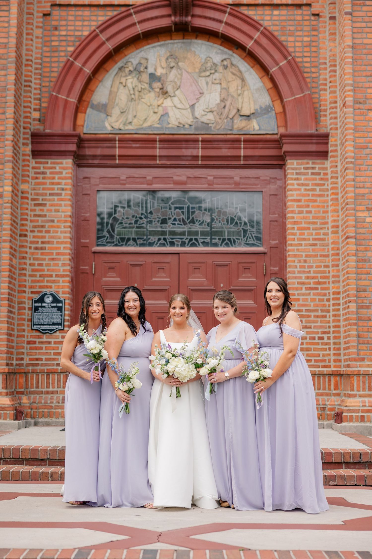 Bride in white gown and four bridesmaids in lavender dresses pose smiling before ornate brick church doors.