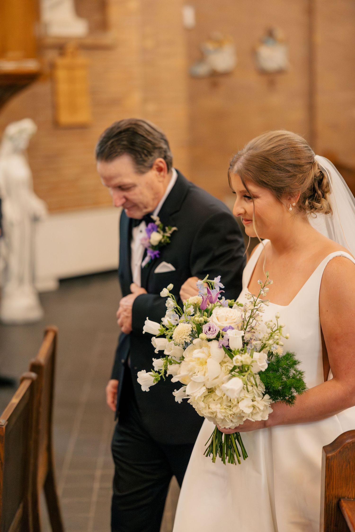 Father escorts smiling bride holding white floral bouquet down the aisle of a Catholic church.