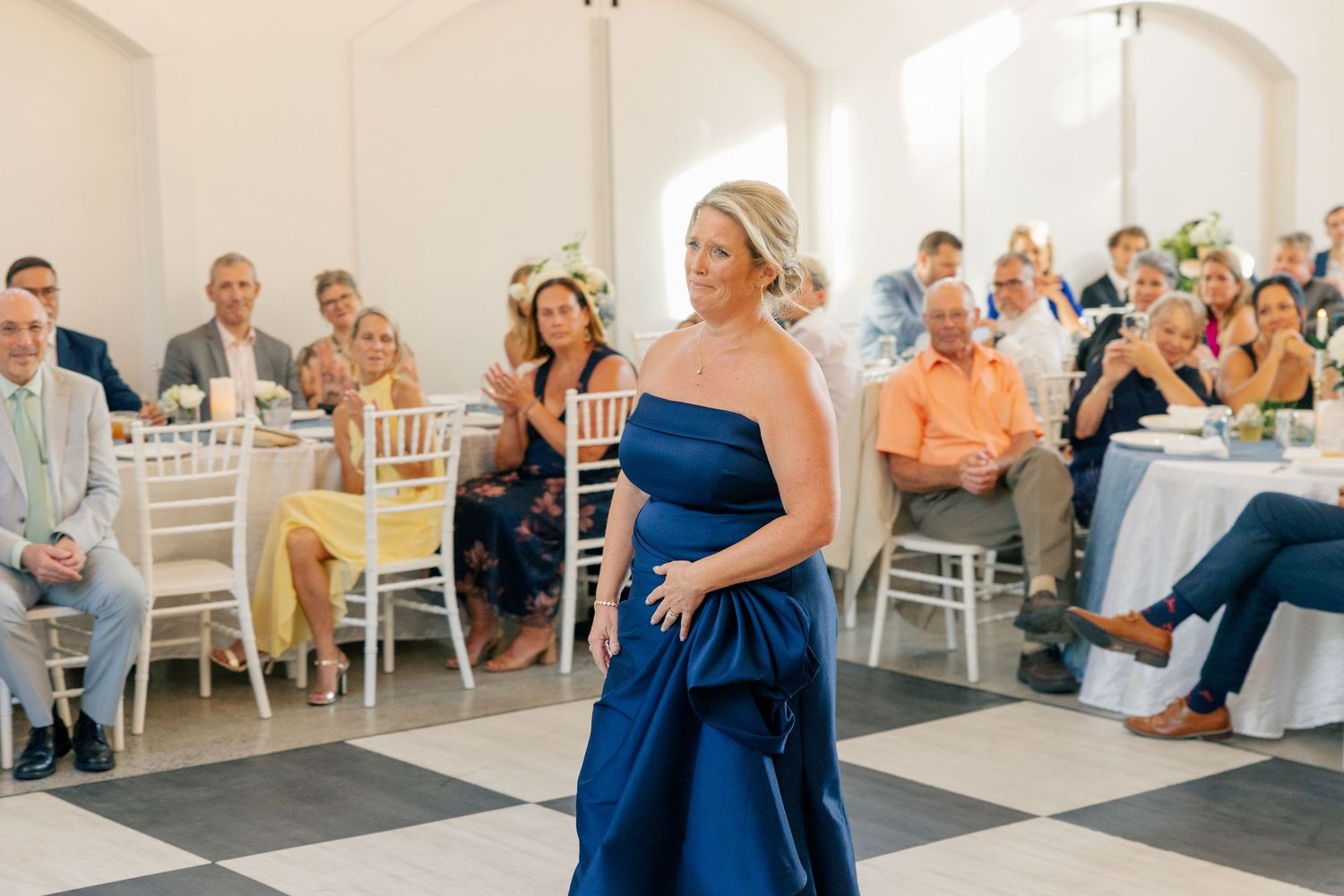 Woman in strapless navy blue gown walks gracefully across checkered dance floor at wedding reception.