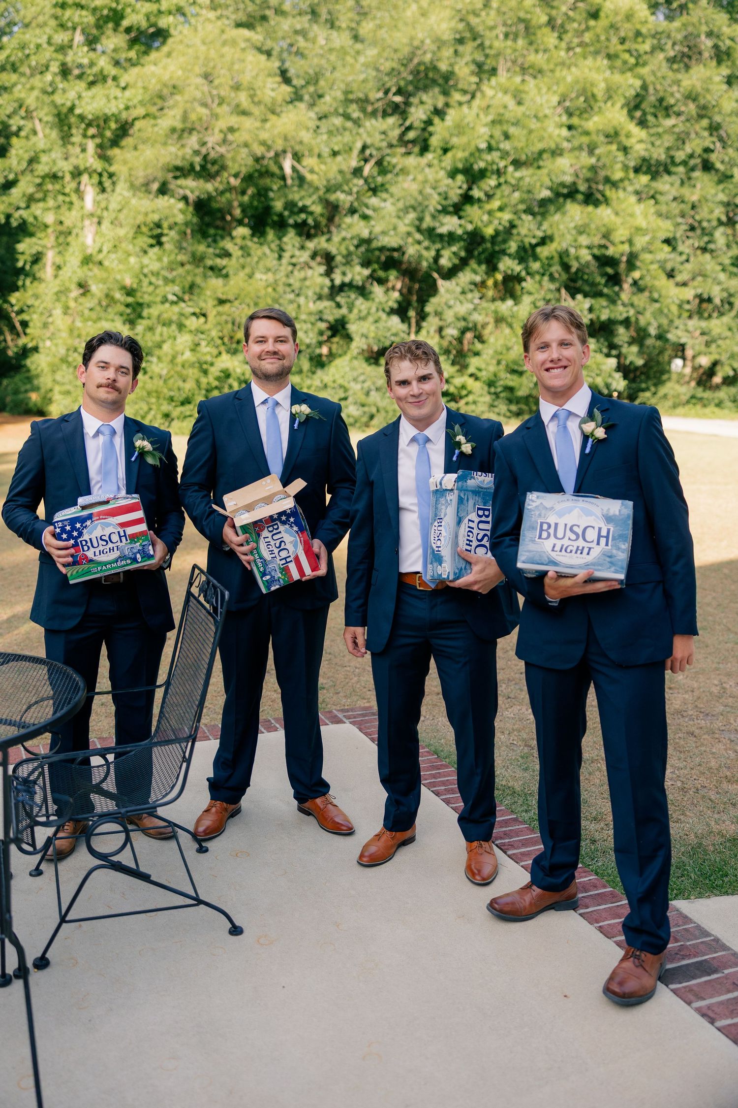 Four groomsmen in navy suits pose outdoors holding cases of Busch Light beer at a wedding reception.