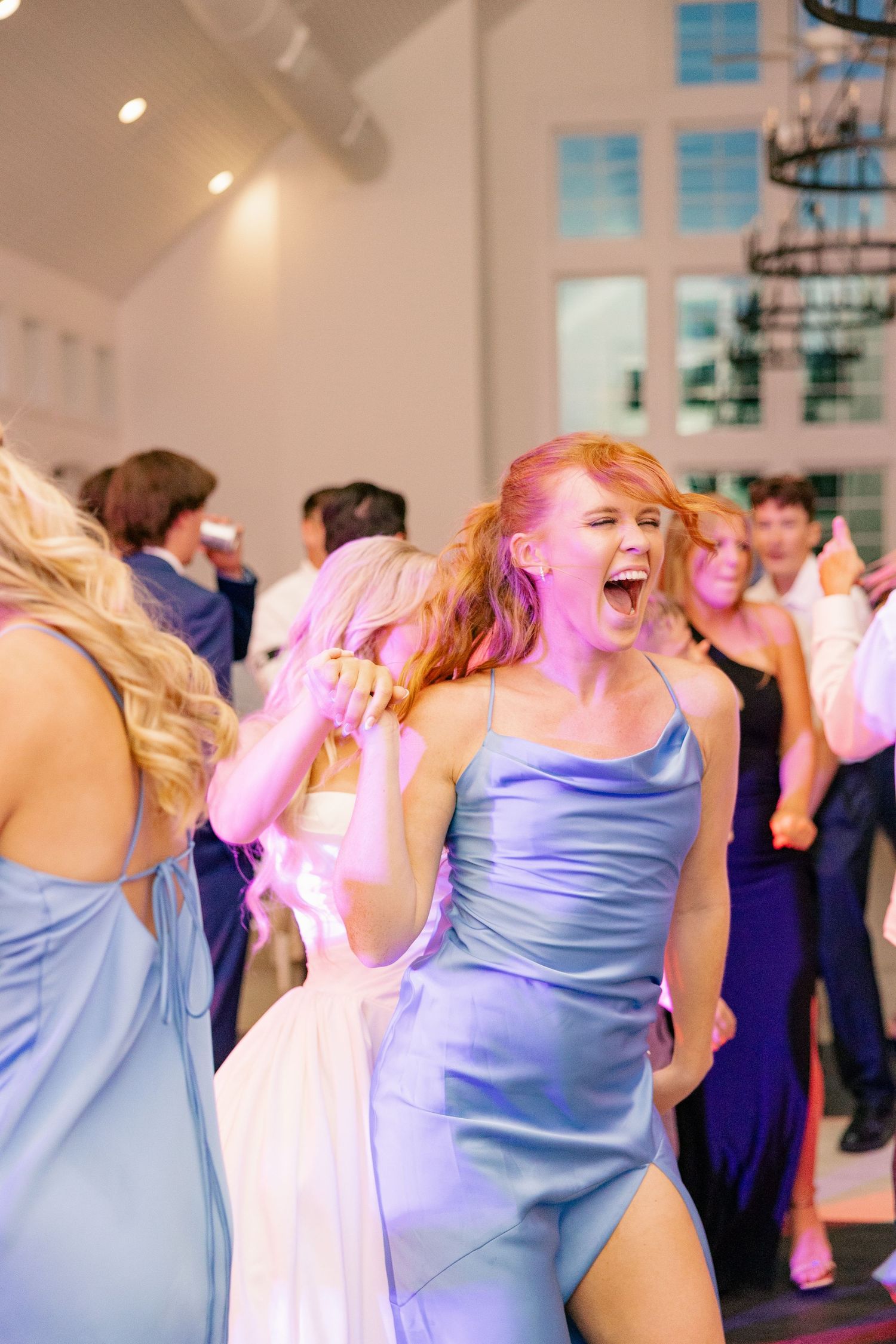 Bridesmaid in blue satin gown dances joyfully at elegant wedding reception with other guests on the dance floor.