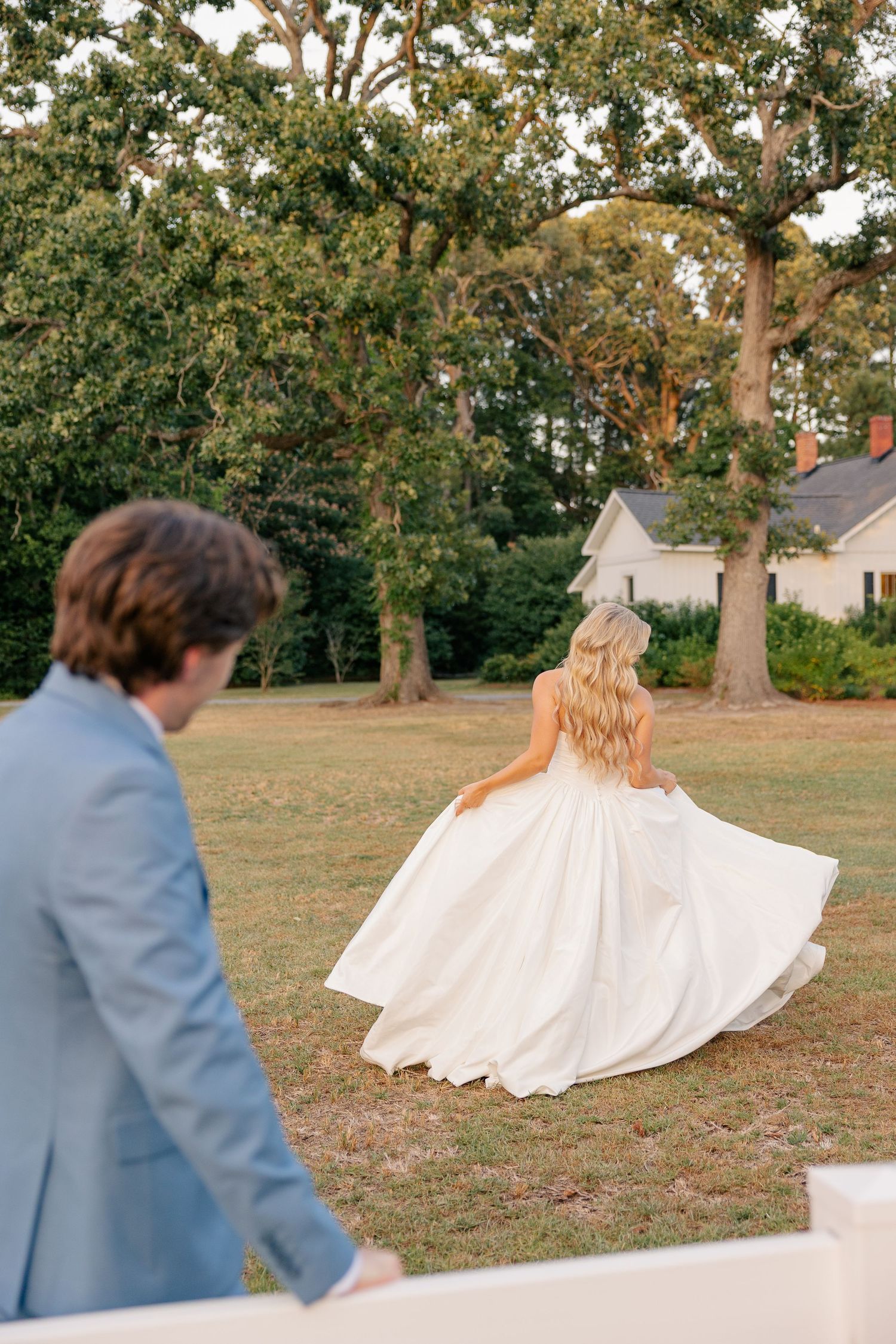 Bride in ballgown twirls on lawn as groom in blue suit watches during golden hour portraits.