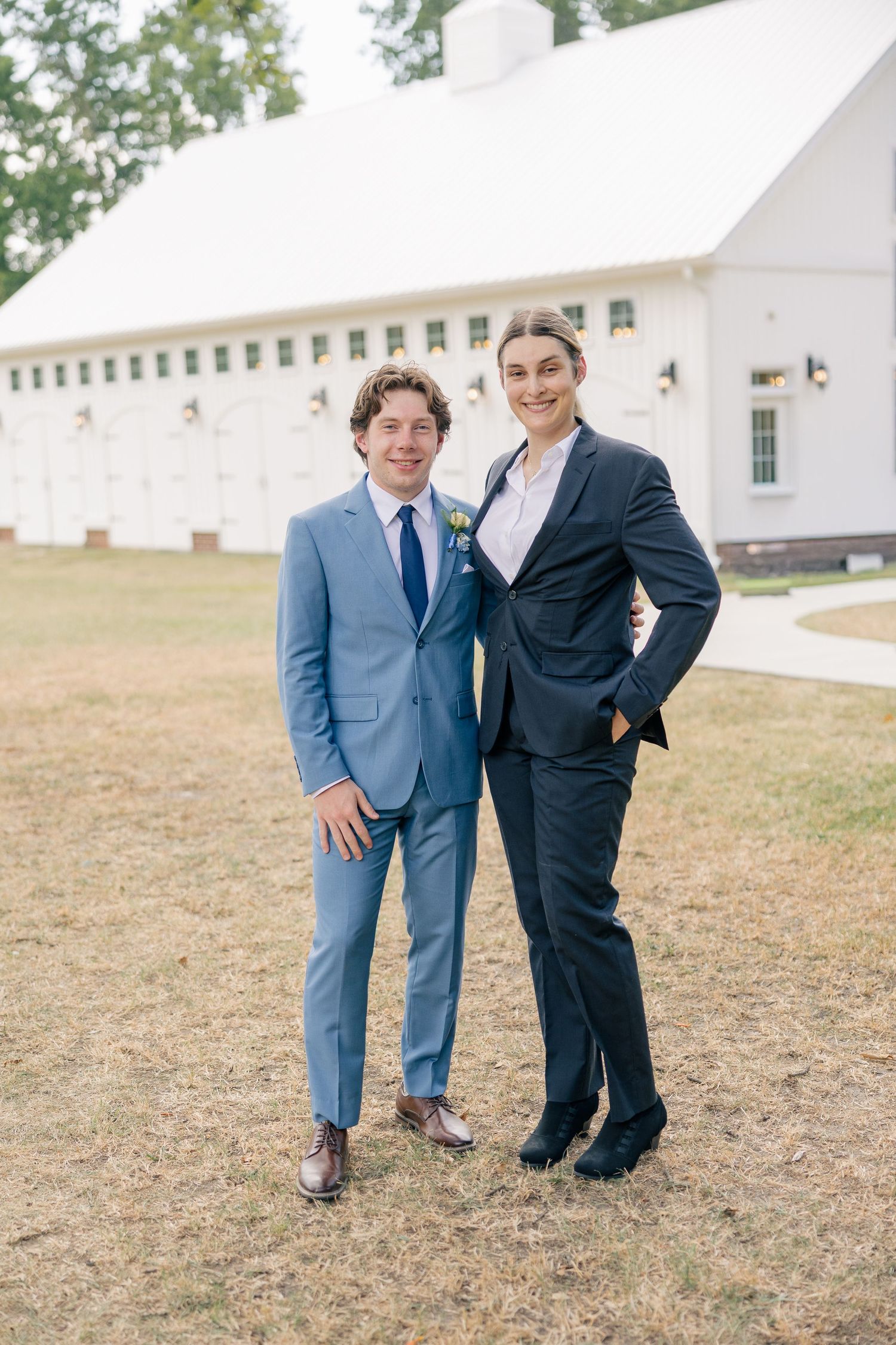 Groom in light blue suit posing with a guest in navy suit outside a white barn venue.