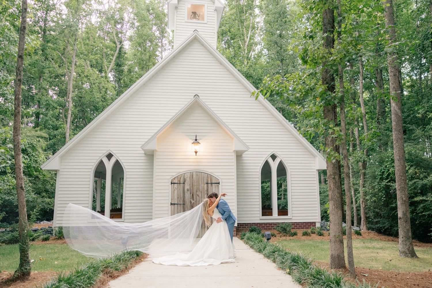 Bride and groom share a romantic kiss outside a white chapel surrounded by lush trees, with veil flowing gracefully.