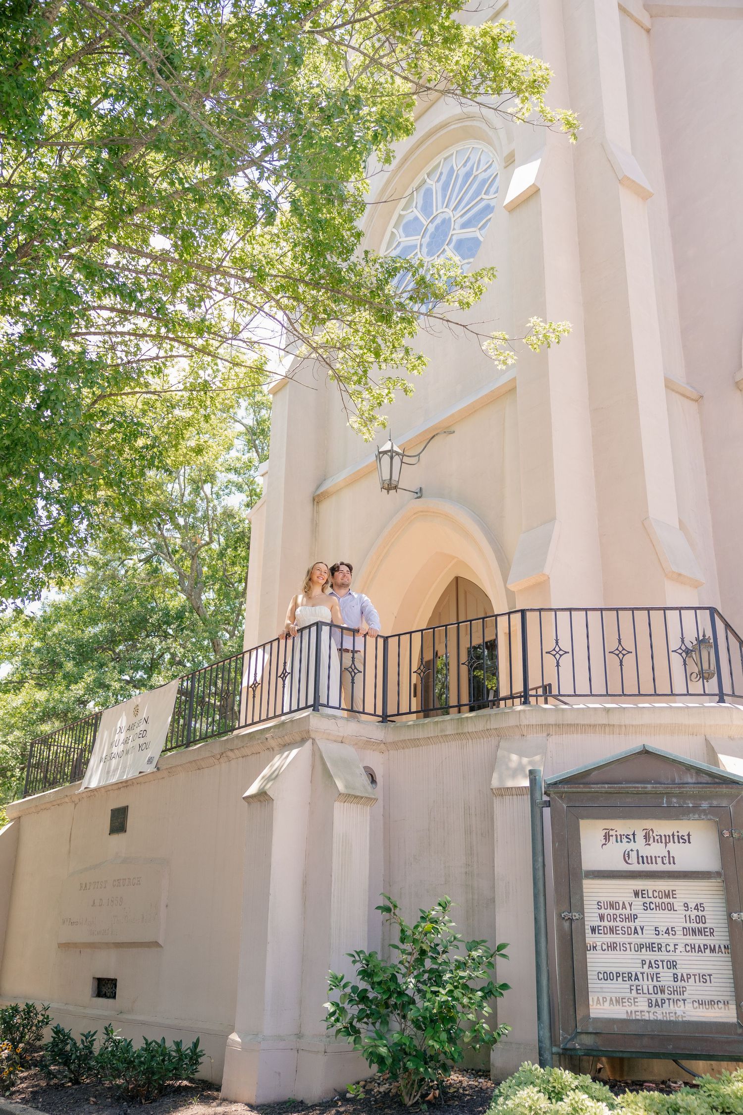 Engaged couple standing on balcony of First Baptist Church surrounded by lush green trees in soft sunlight.