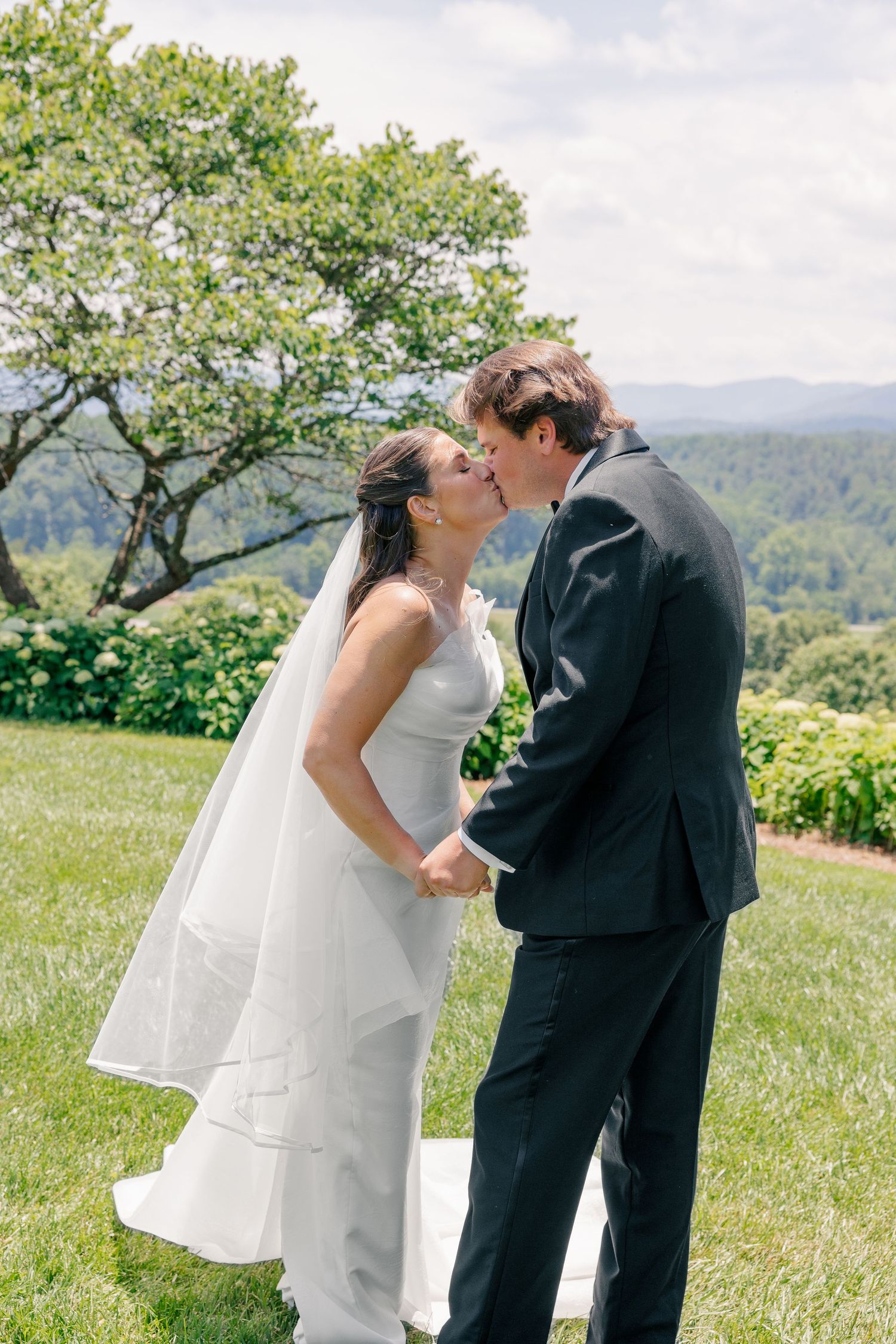 Bride and groom share first kiss outdoors with scenic mountain views behind them.