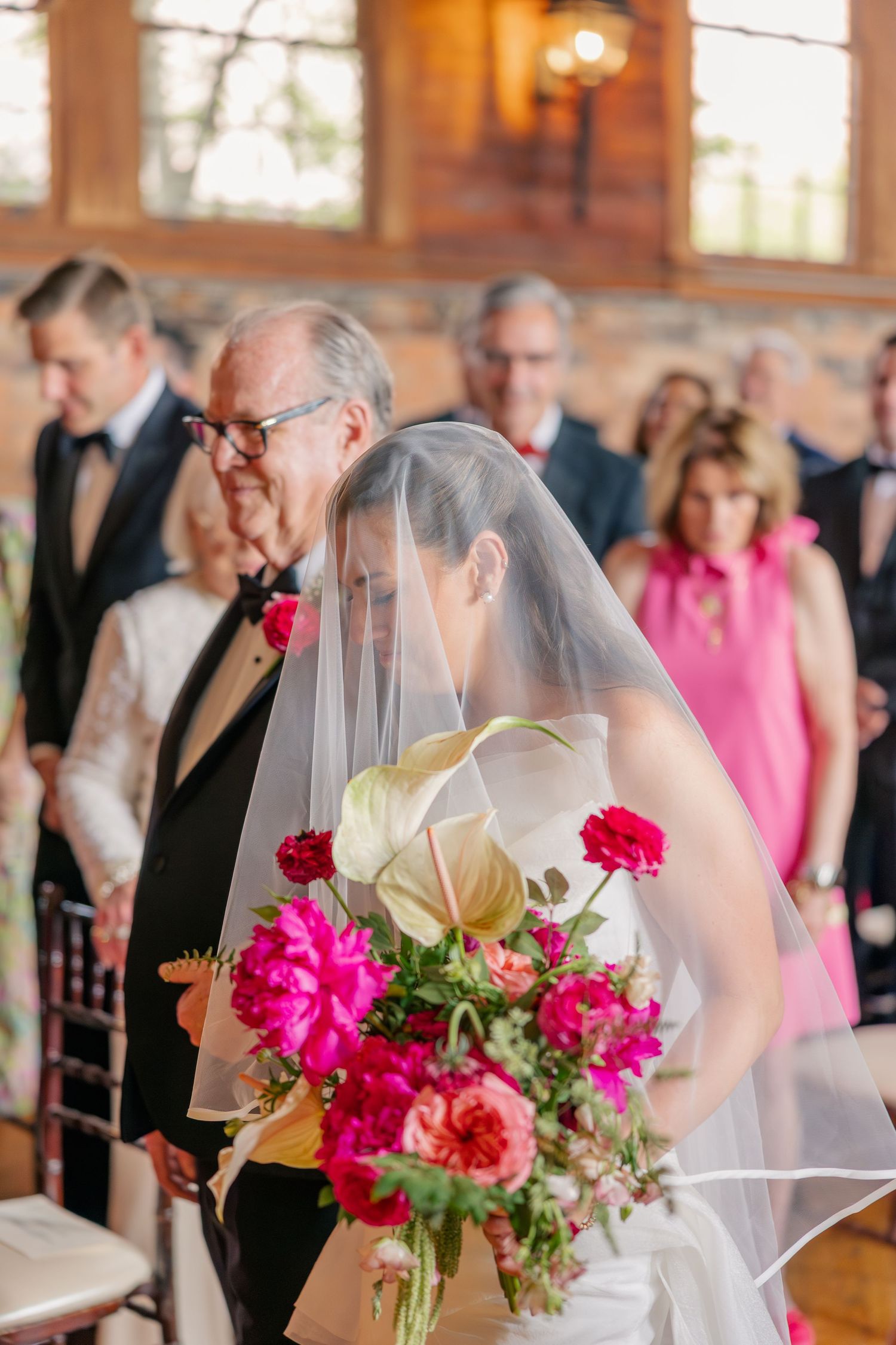 Bride in white strapless gown and veil walking down aisle with father holding bright pink bouquet.