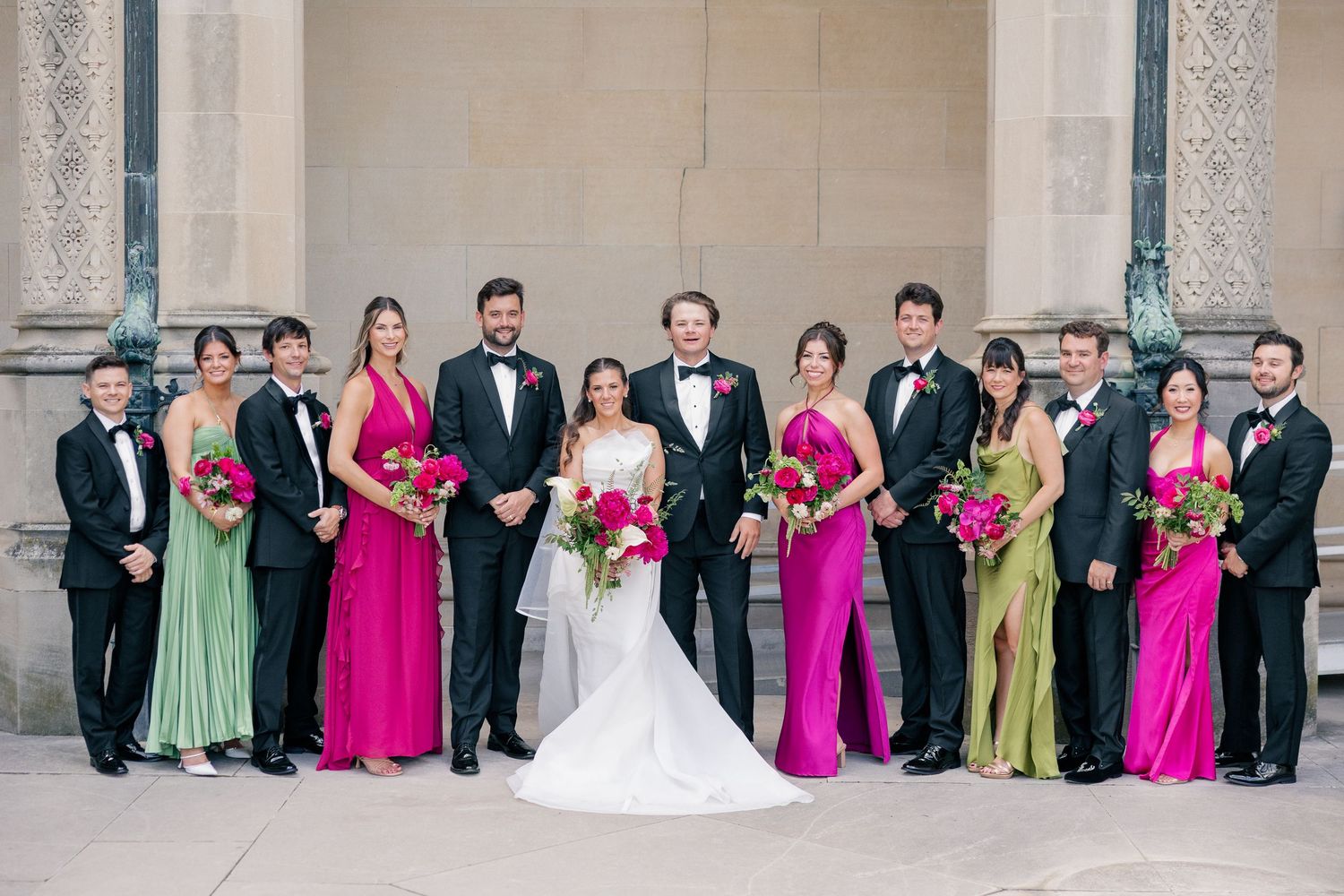 Wedding party in fuchsia and green gowns with groom and groomsmen in black tuxedos outdoors.