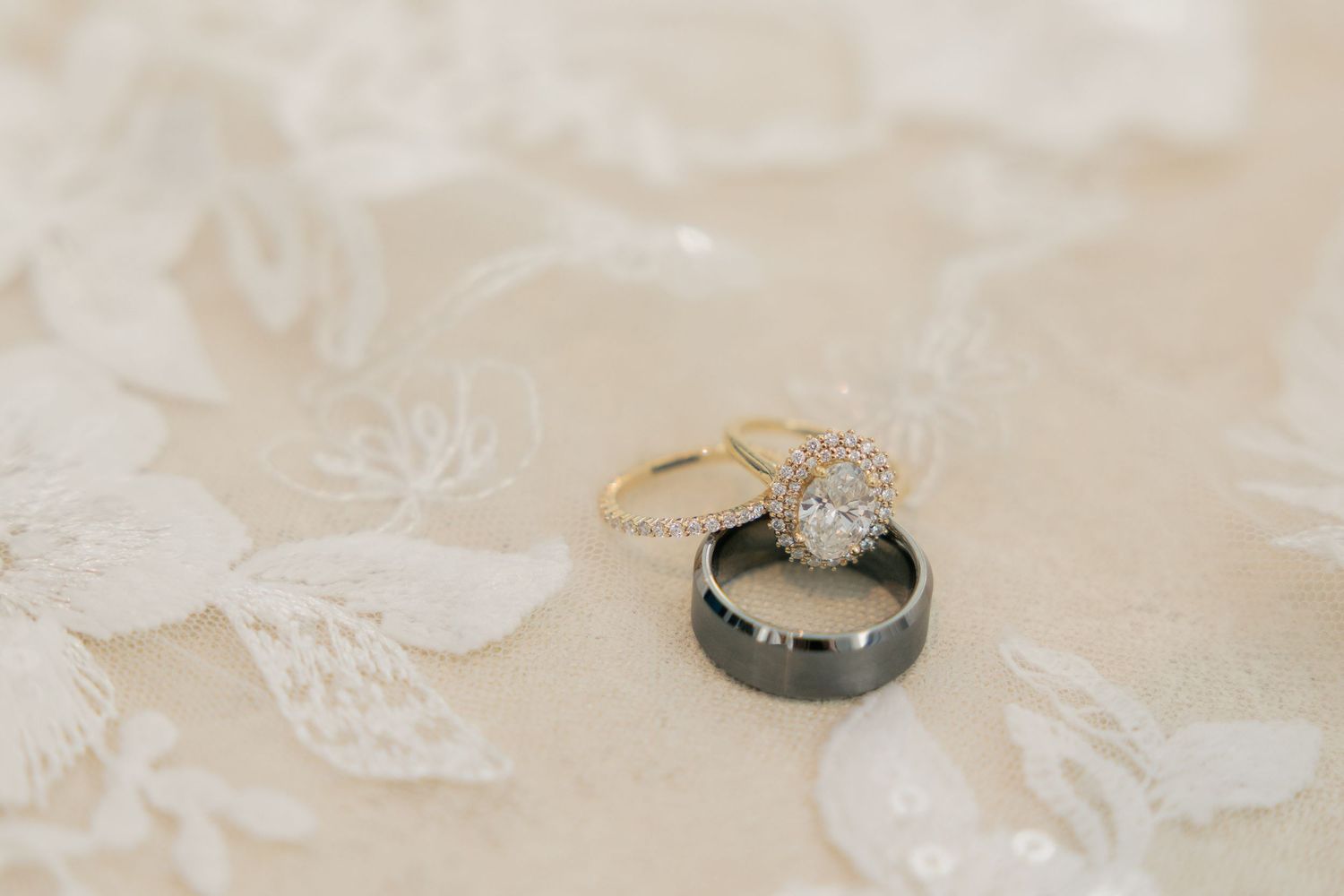 Gold halo diamond engagement ring stacked with a dark tungsten wedding band on white lace fabric.