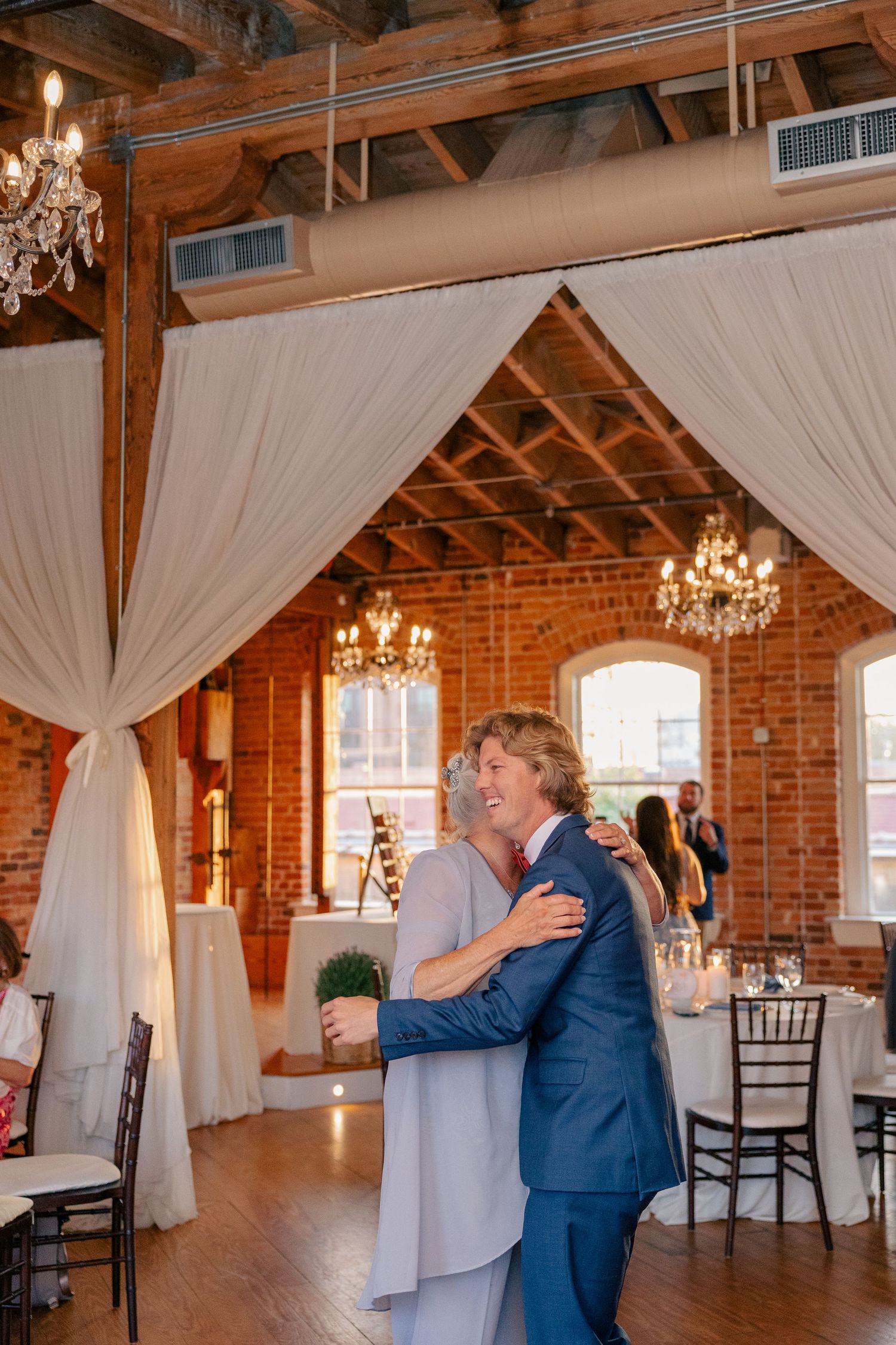 Groom shares joyful mother-son dance in elegant rustic brick reception hall adorned with chandeliers and draping.