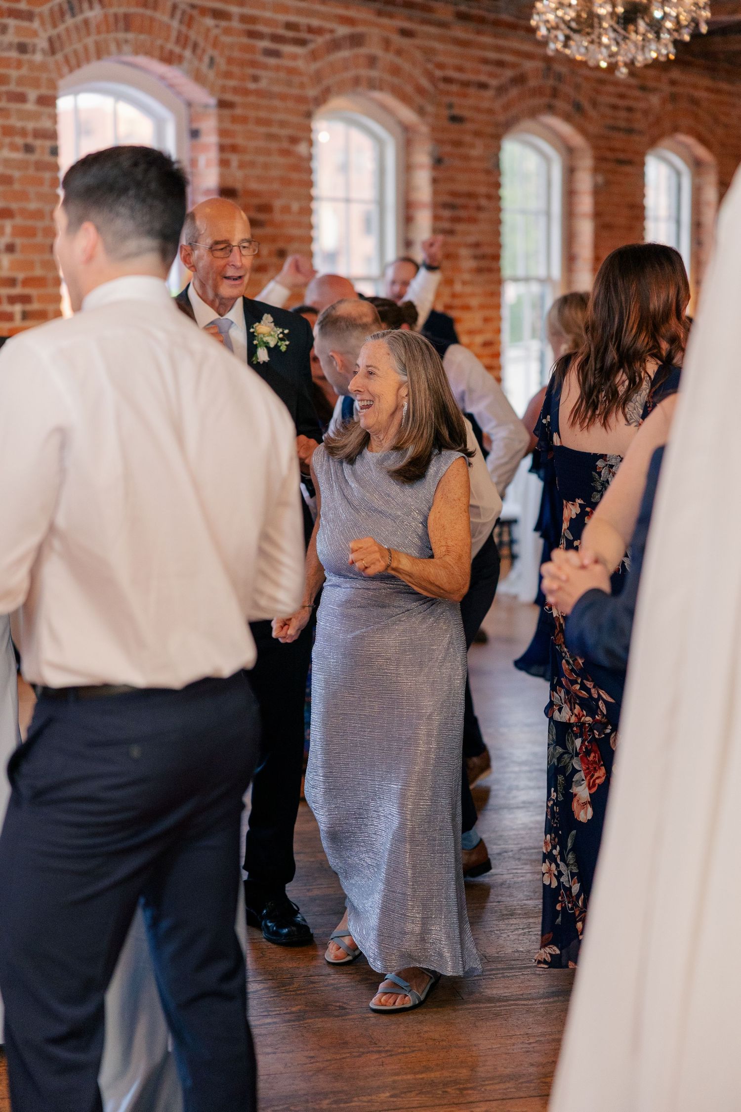 Guests smile and dance together on the wooden dance floor during a wedding reception in a brick ballroom.