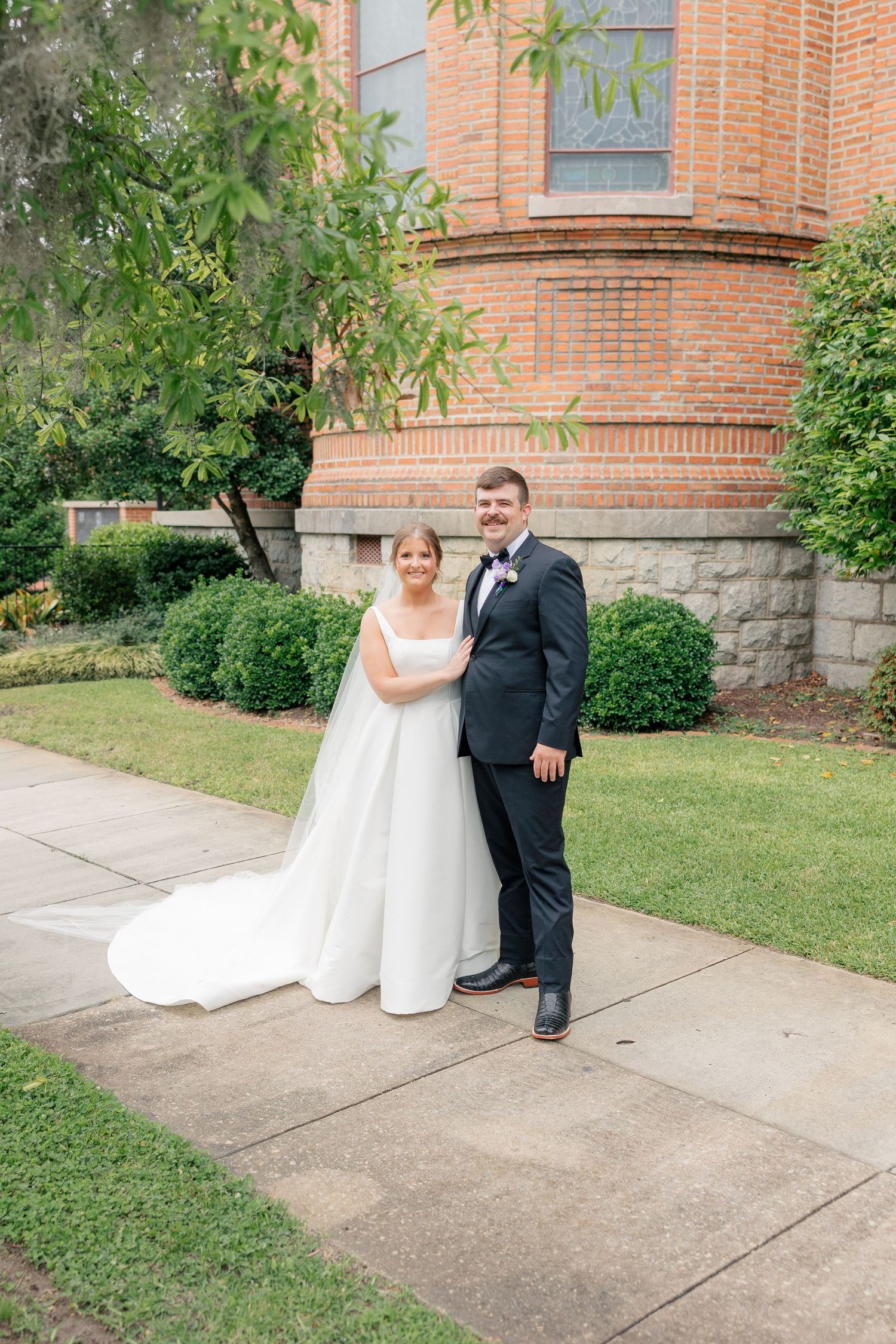 Bride in white gown with cathedral veil and groom in black tuxedo pose outside brick church.