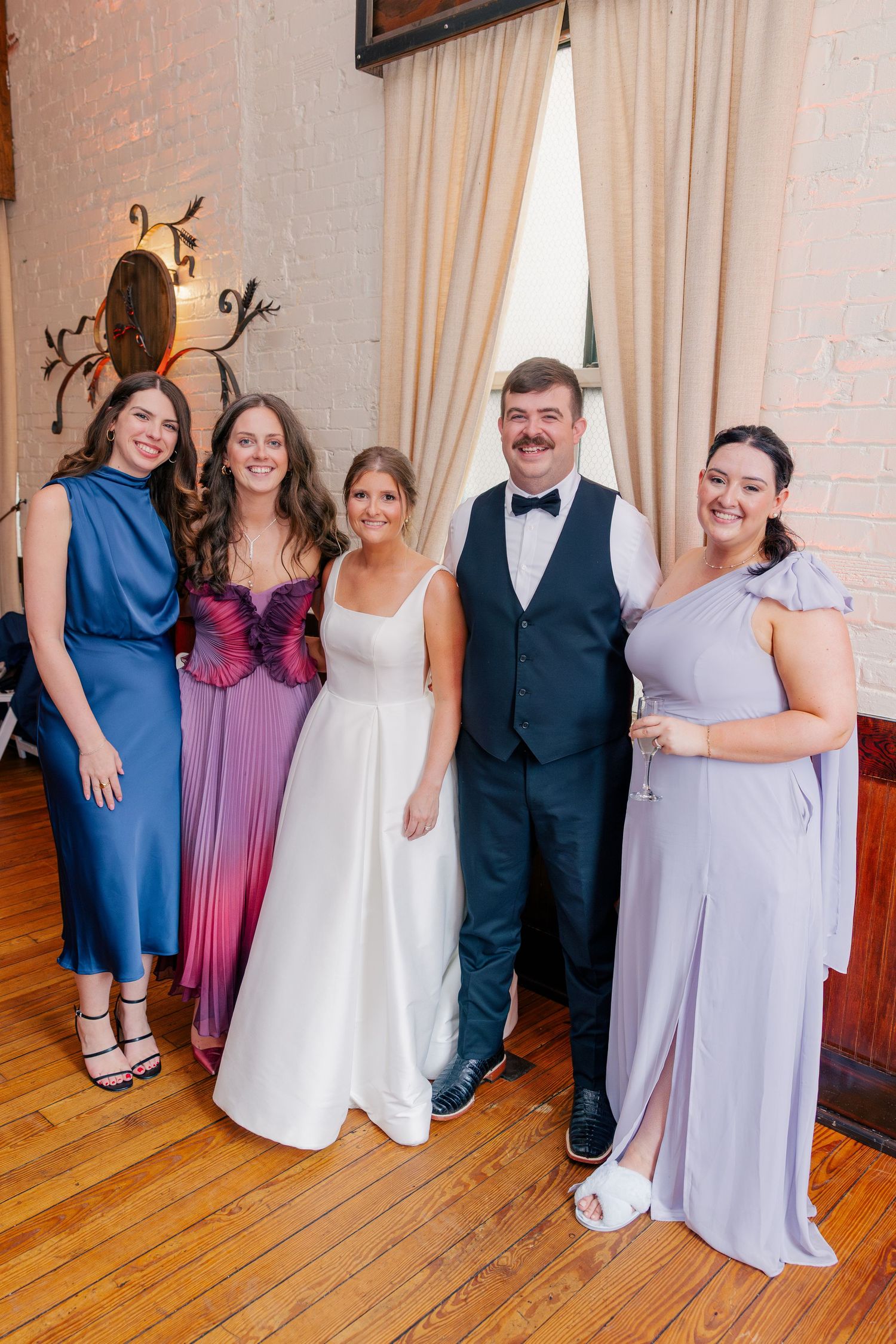 Wedding party posing together, bride in white gown with groom in tuxedo and bridesmaids in colorful formal dresses.