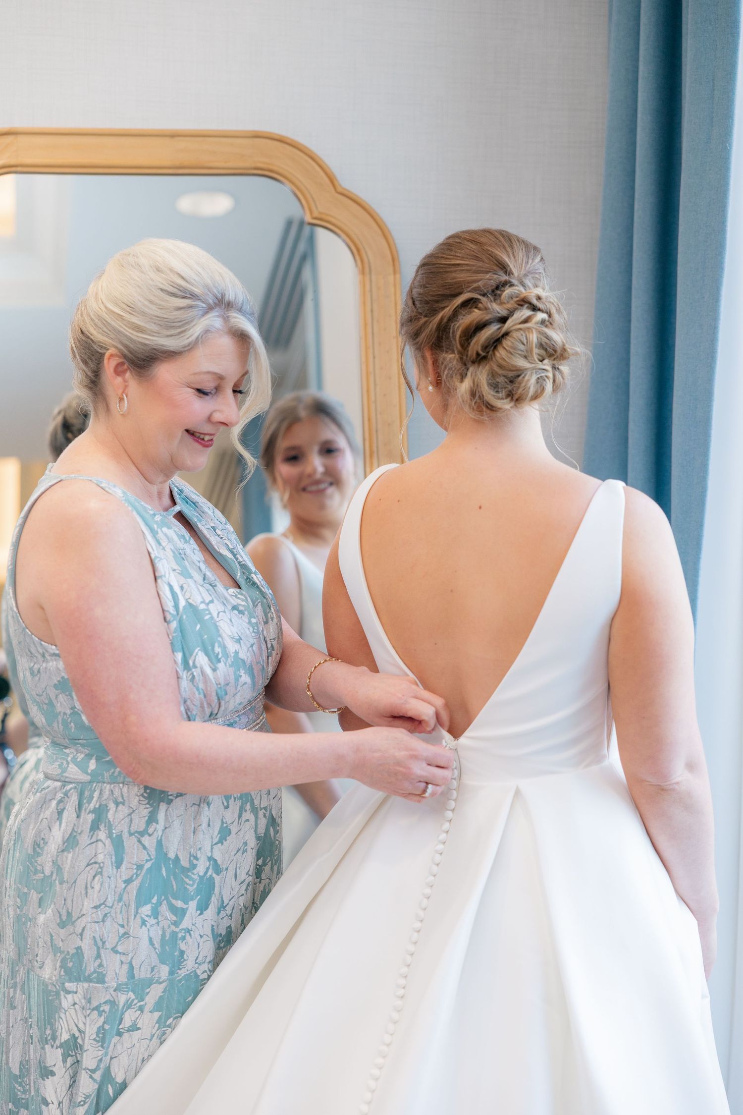 Mother of bride fastens buttons on back of bride's white gown with deep V-back before ceremony.