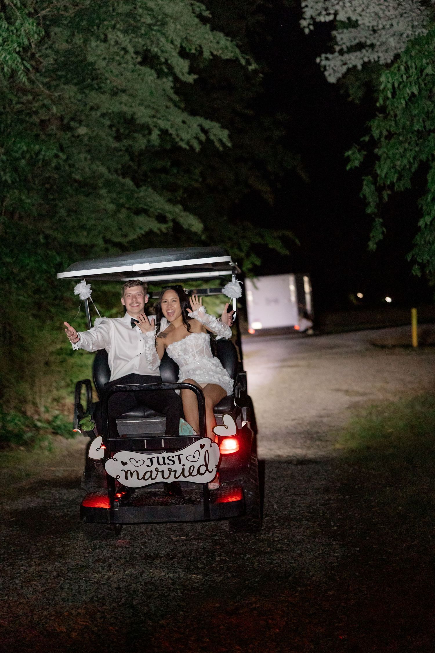 Newlyweds ride away in a car decorated with 'Just Married' sign on a dark wooded path.