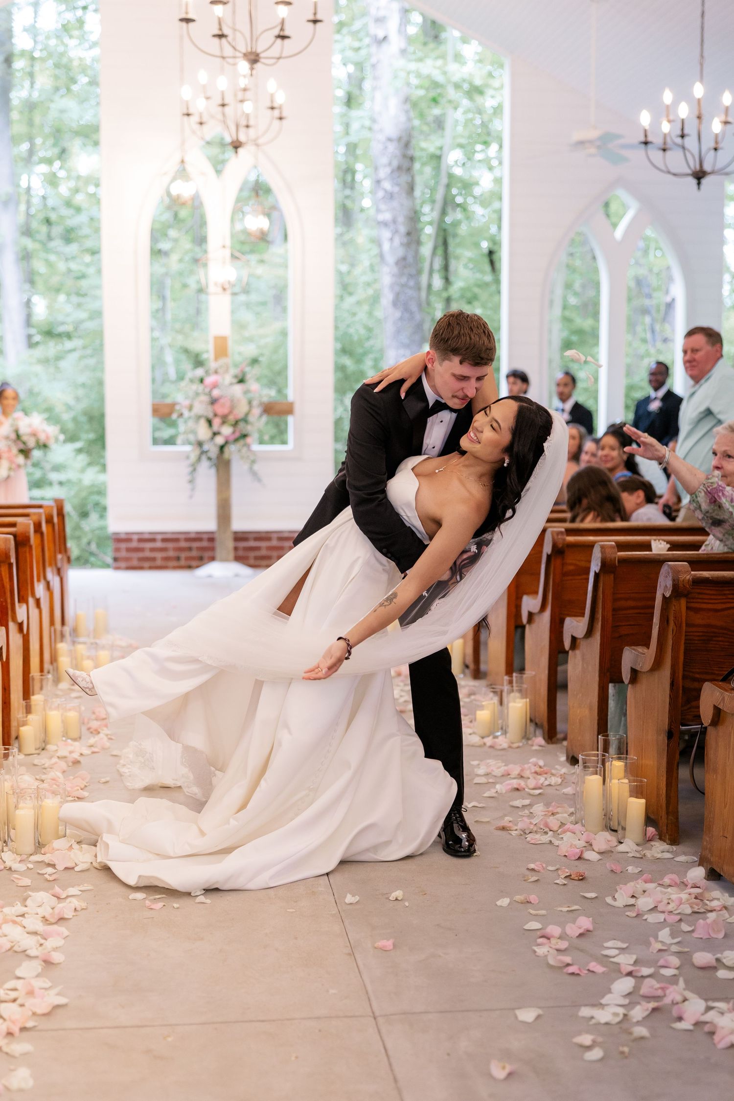 Romantic dip pose by newlyweds in white chapel with flower petals and wooden pews.