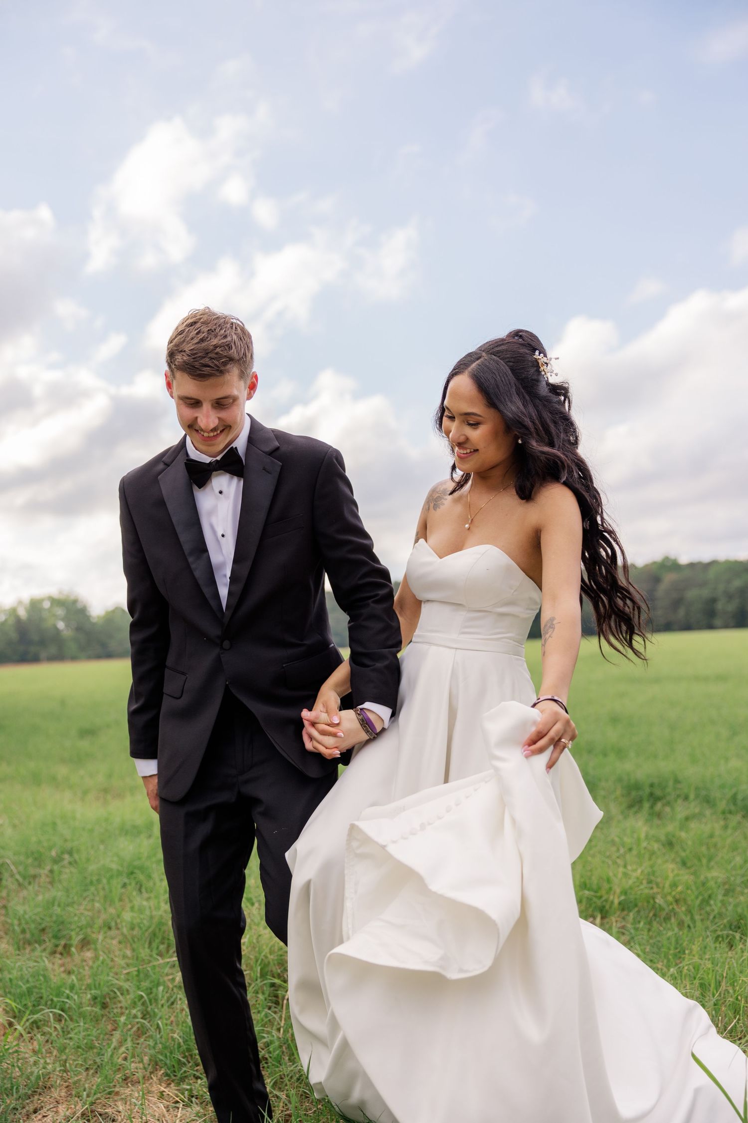 Bride and groom walking hand in hand through grassy field under cloudy sky.