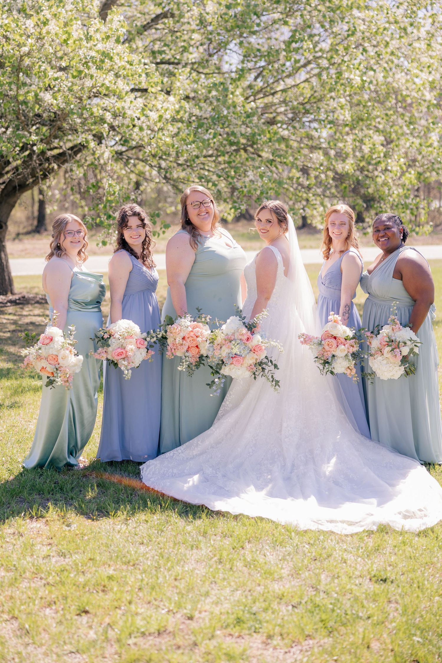 Bridesmaids in soft blue dresses hold pastel bouquets while posing together on grass in sunlight.