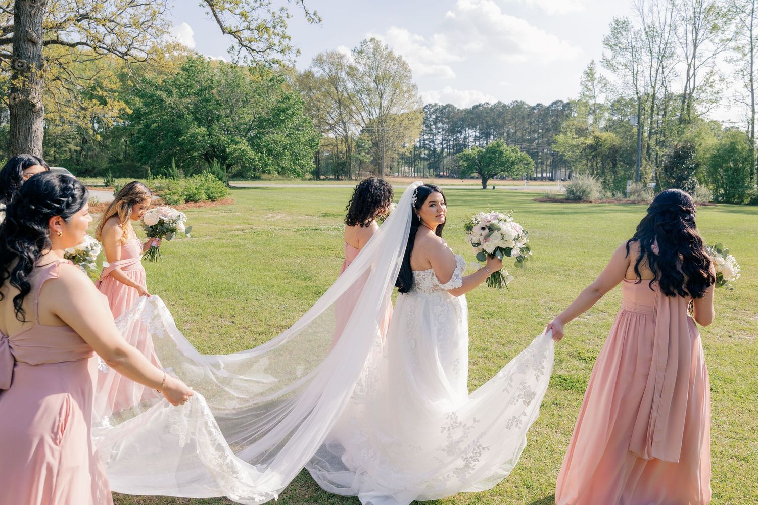 Bridesmaids in pink dresses help adjust the bride's flowing wedding gown train outdoors.