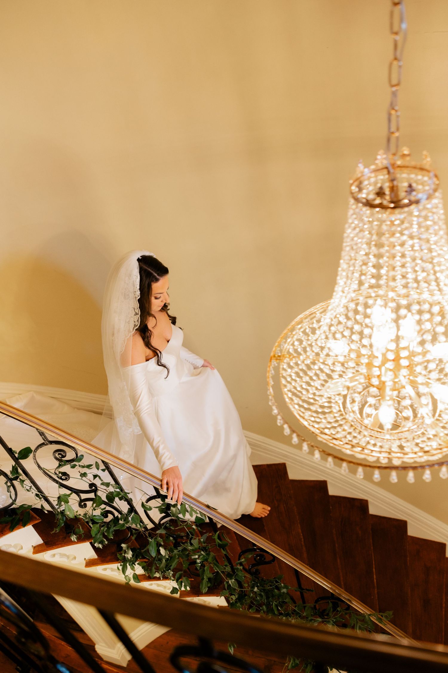 A bride in a white gown sits gracefully on elegant stairs beneath a crystal chandelier.