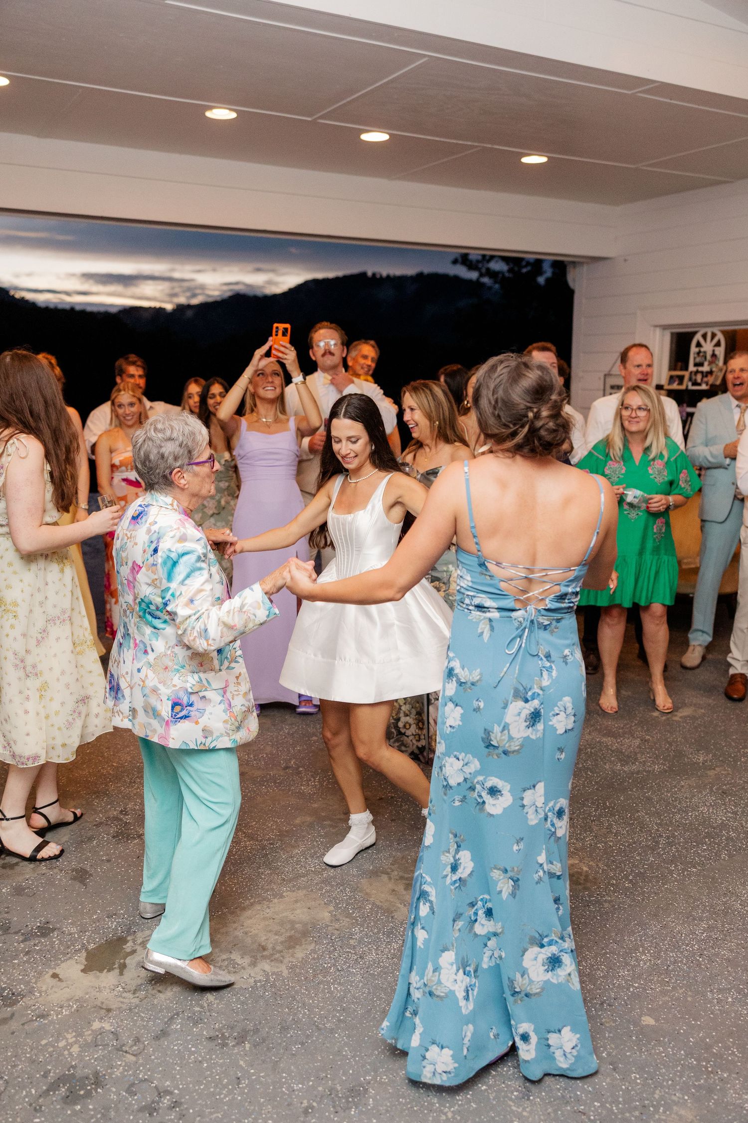 Wedding guests dance together in floral and pastel dresses during a lively reception celebration.