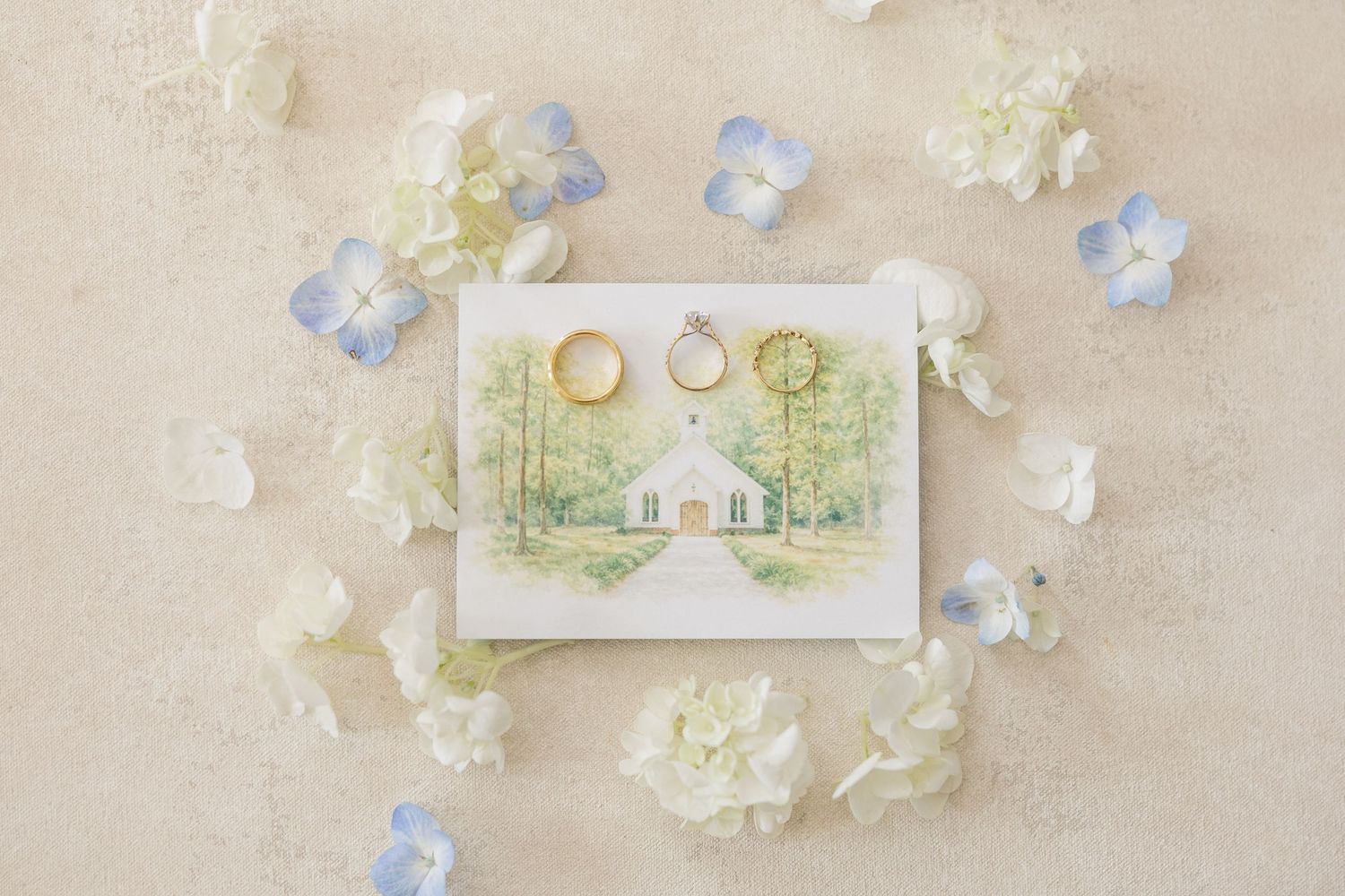 Wedding rings and watercolor chapel illustration surrounded by blue and white hydrangea petals.