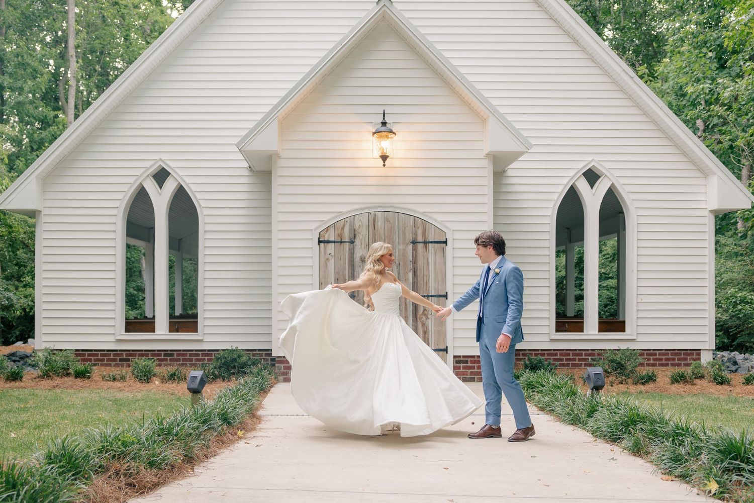 Bride and groom dancing in front of a white chapel surrounded by lush greenery on their wedding day.