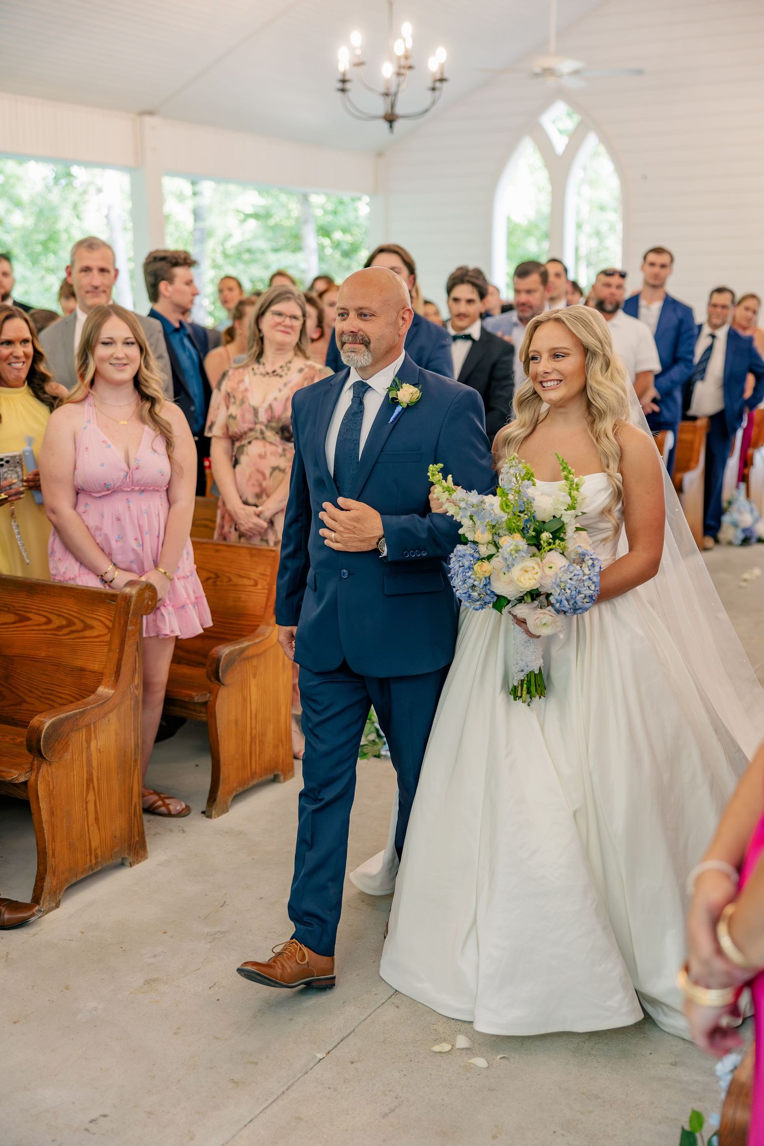 Father proudly escorts radiant bride down aisle of white chapel filled with wedding guests.
