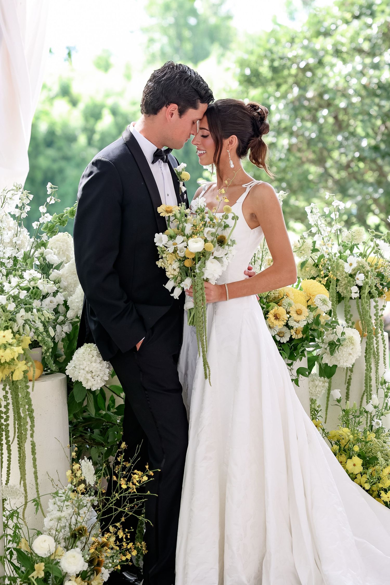 Bride in white gown holding yellow and white bouquet stands with groom in black tuxedo amid lush floral arrangements.