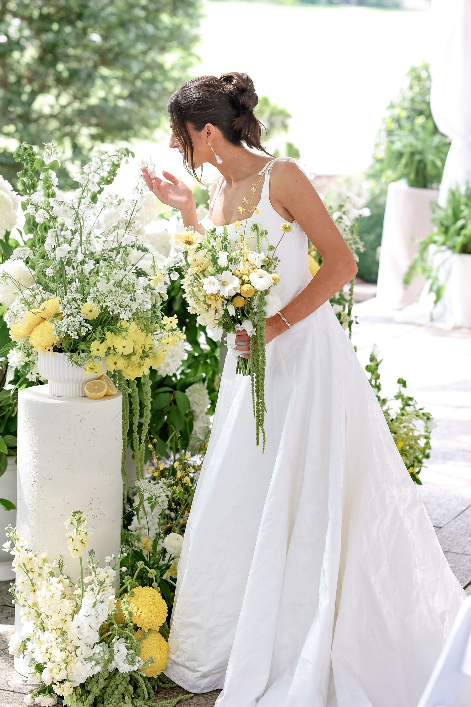 Bride in white gown holds yellow and white bouquet beside floral arrangement with lemons at outdoor wedding.
