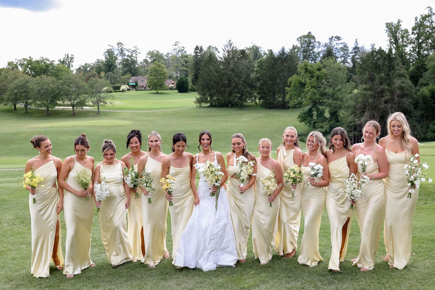 Bride in white gown poses with bridesmaids in yellow dresses holding bouquets on lush green golf course.