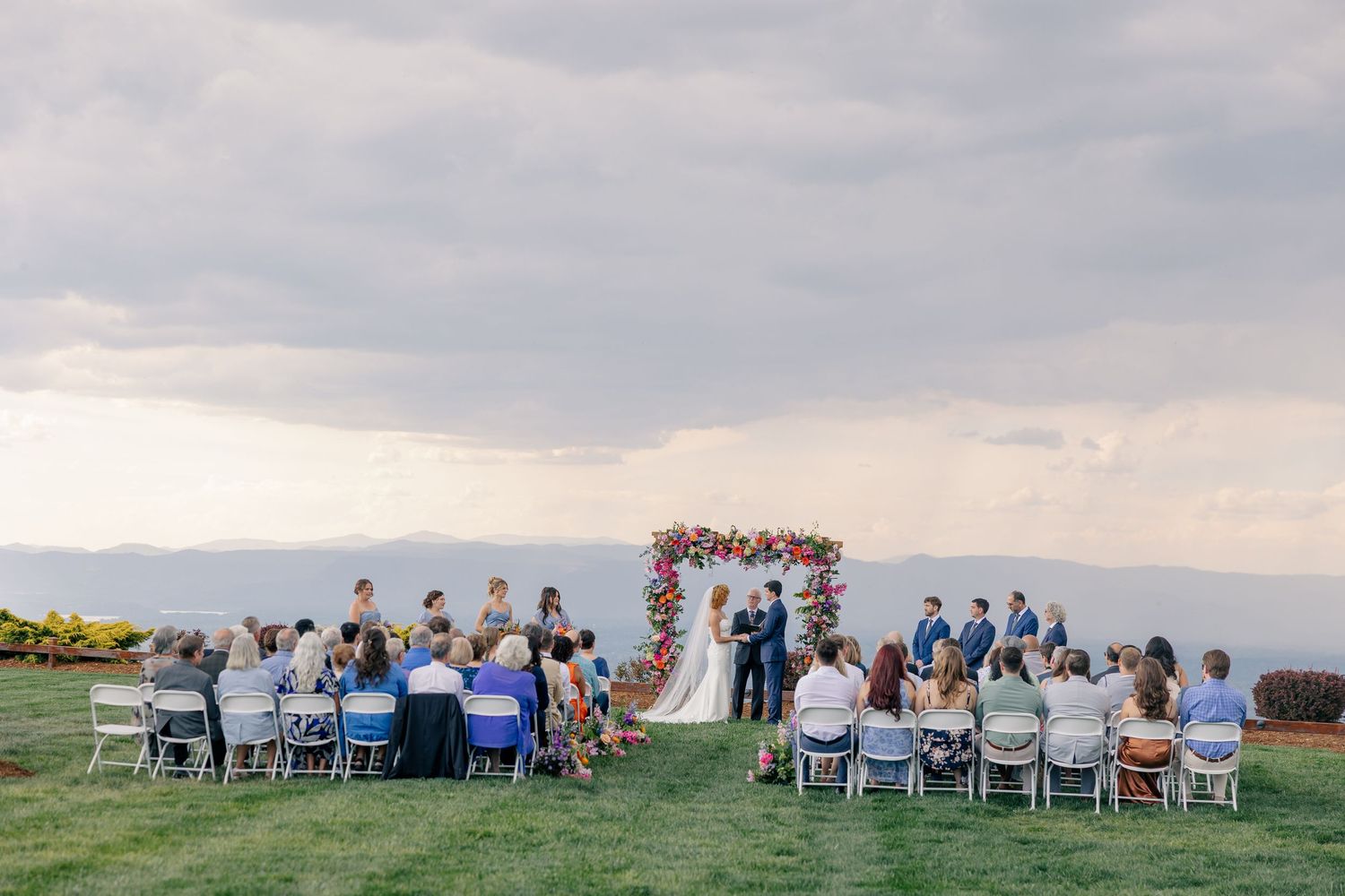 Outdoor wedding ceremony on a hilltop with floral arch, guests seated on white chairs, and a scenic cloudy sky backdrop.