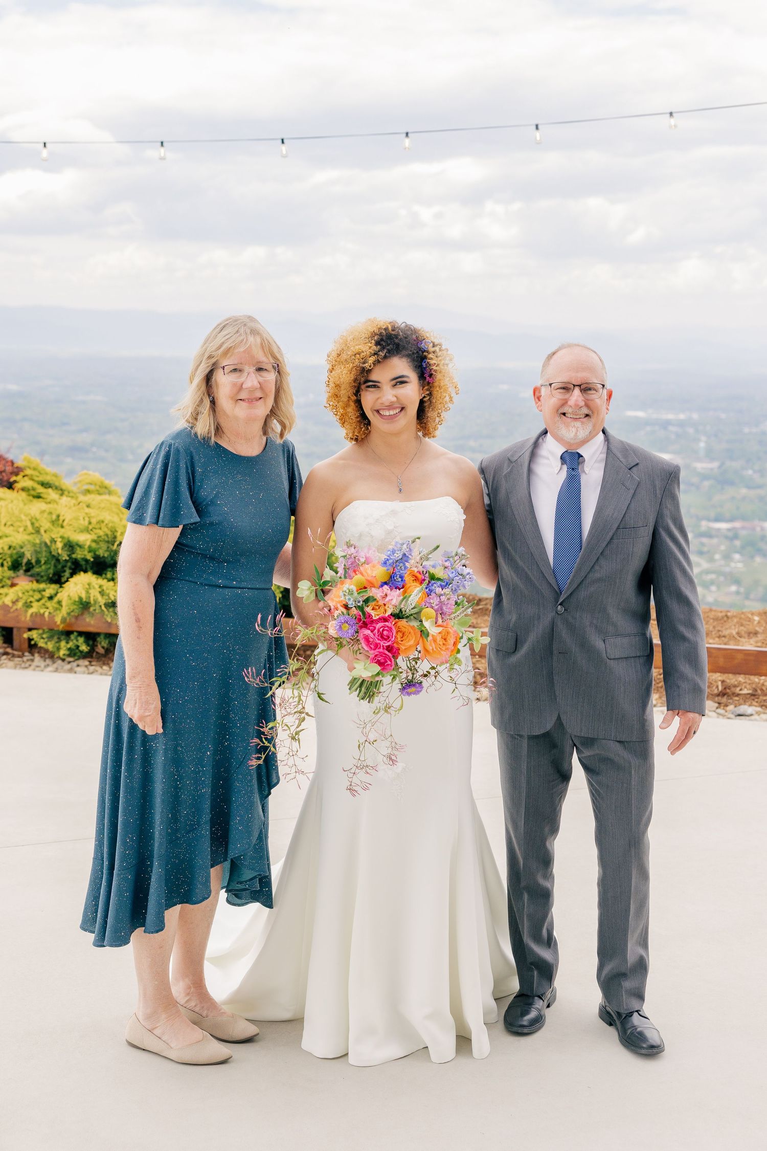 Bride holding colorful bouquet posing with parents on scenic overlook wedding venue.