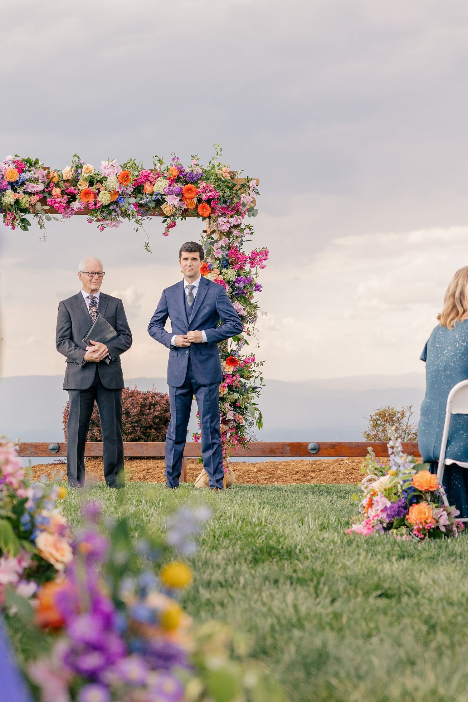 Groom in navy suit awaiting bride at outdoor ceremony beneath a colorful floral arch with mountain views.