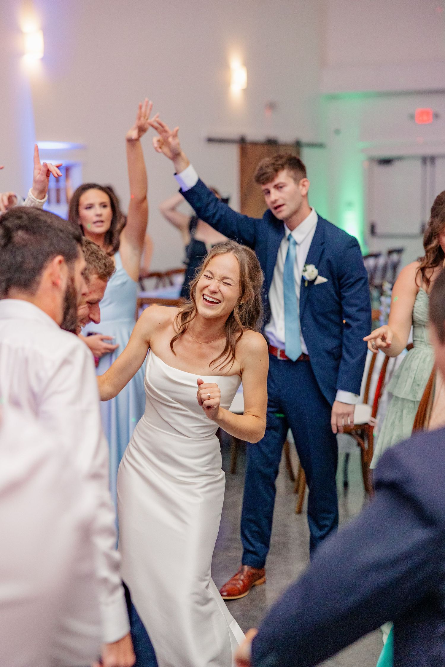 Bride in white gown laughing and dancing with guests at lively indoor wedding reception.